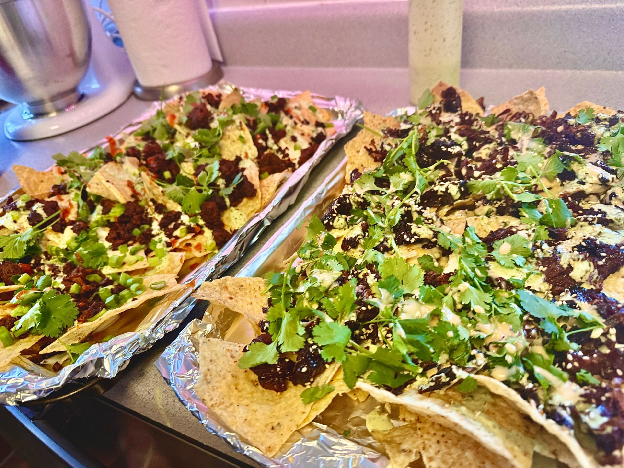Two large nachos topped with green cilantro, black sauce, and cheese, served on foil-lined trays with condiments in the background.