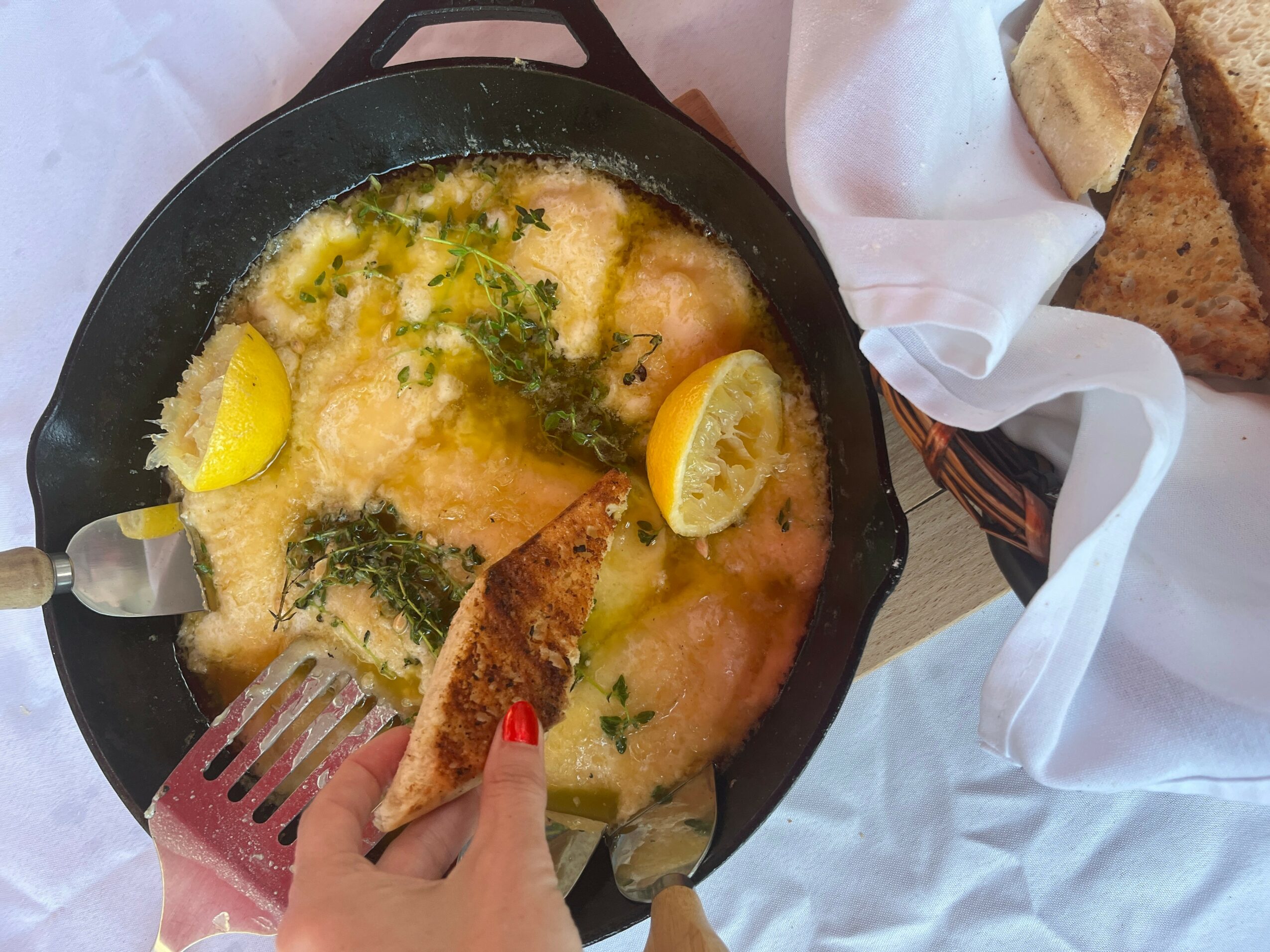 Person holding a piece of toasted bread over a skillet with cooked fish, lemon wedges, and herbs, on a white tablecloth.