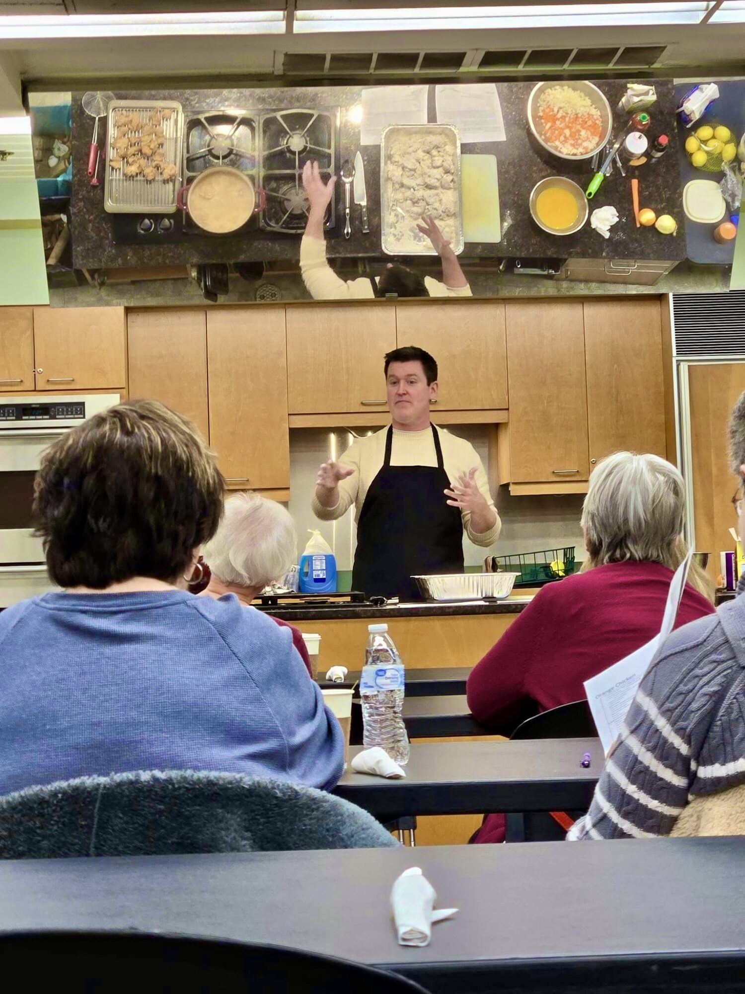 Cooking class with instructor in apron teaching students in a kitchen classroom, viewed from above, with ingredients and utensils on the counter.