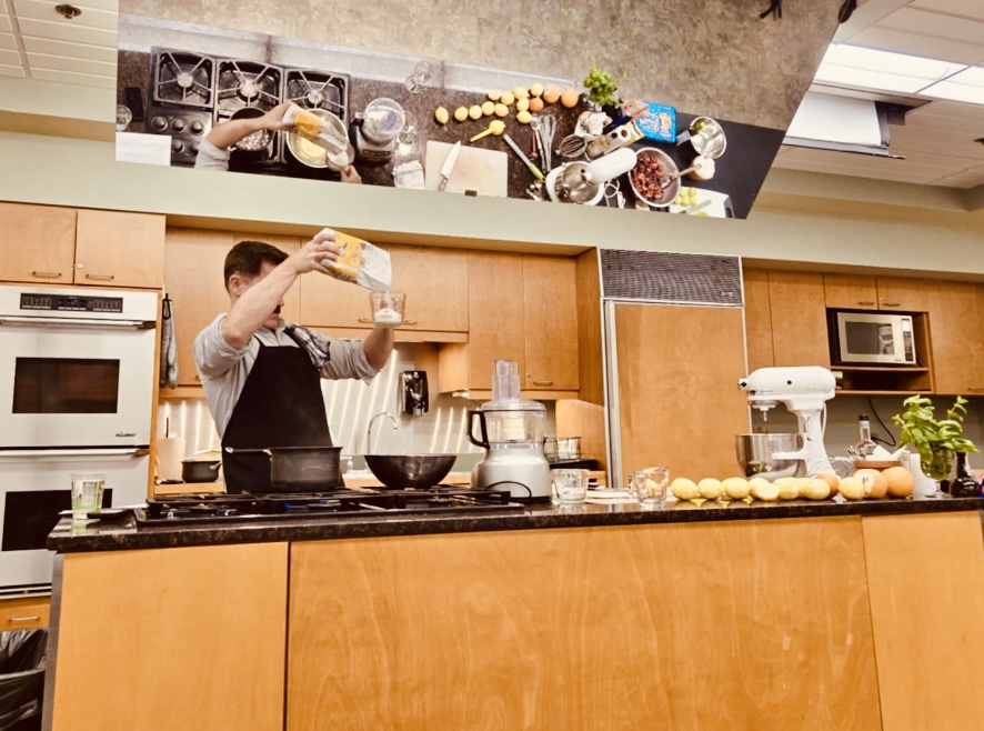 Person in a kitchen pouring liquid into a container, with kitchen appliances and ingredients on the counter.