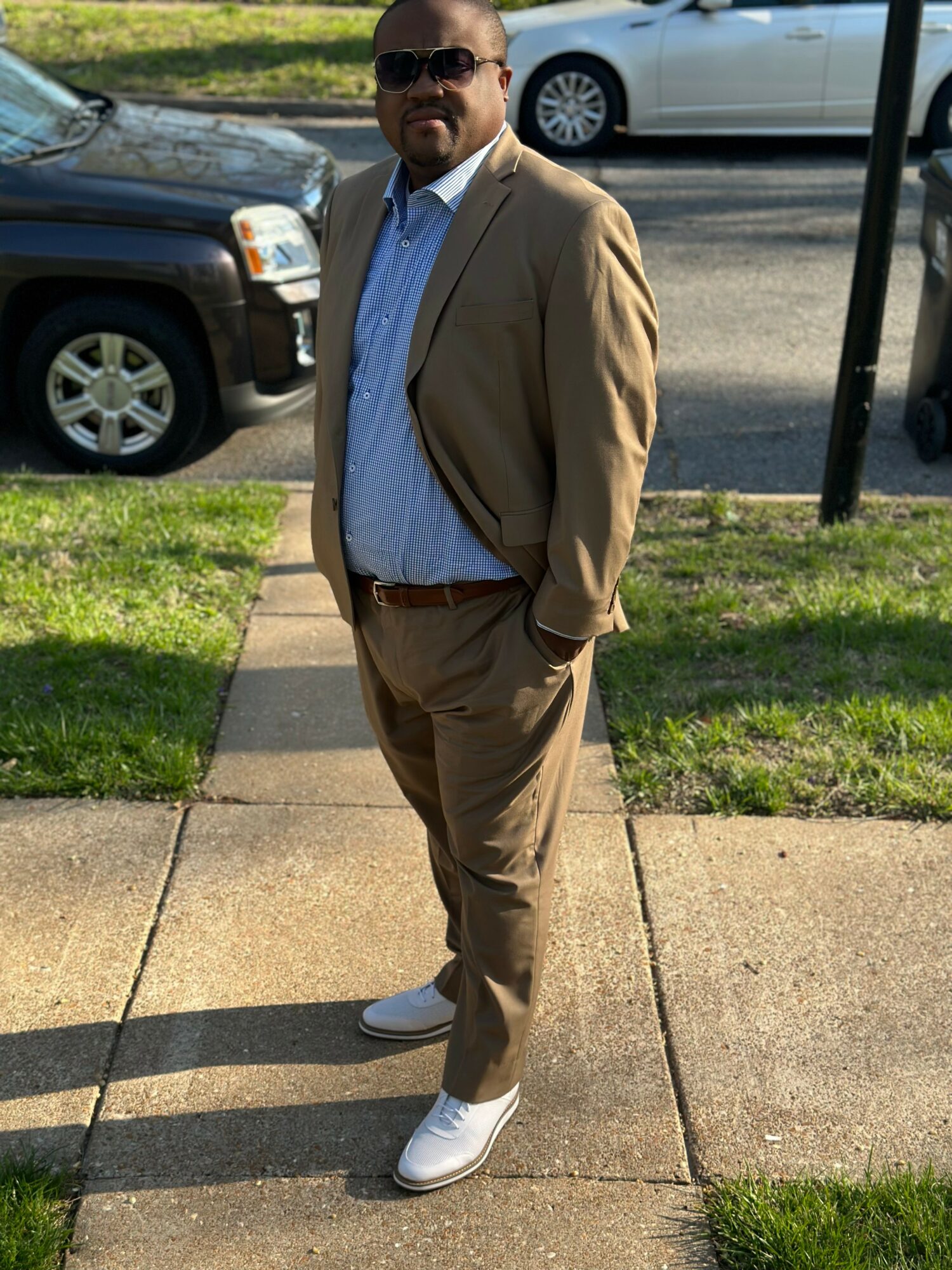 Man in beige suit and sunglasses standing on sidewalk with cars in background.