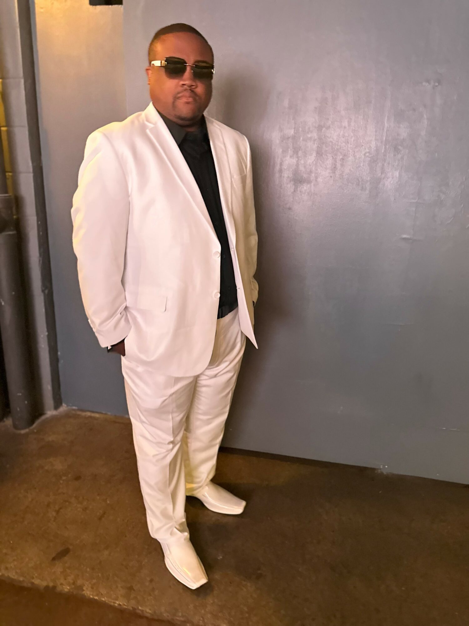 Man in a white suit with sunglasses standing against a plain wall, hands in pockets, looking at the camera.