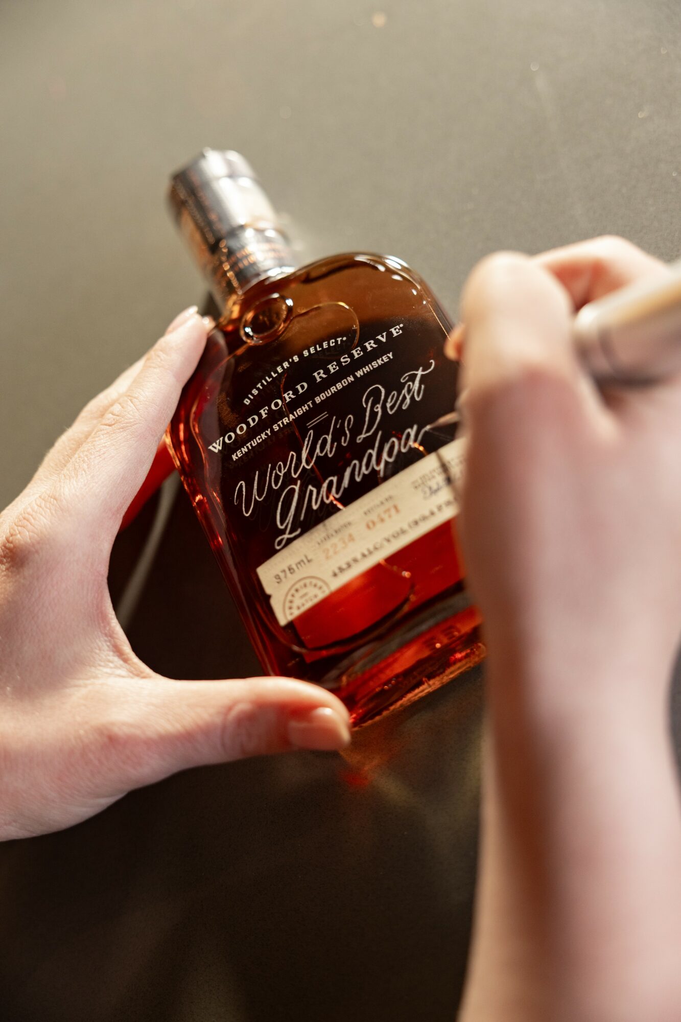 Person writing on a bottle labeled 'World's Best Grandma' with a pen, held in hands over a dark surface.