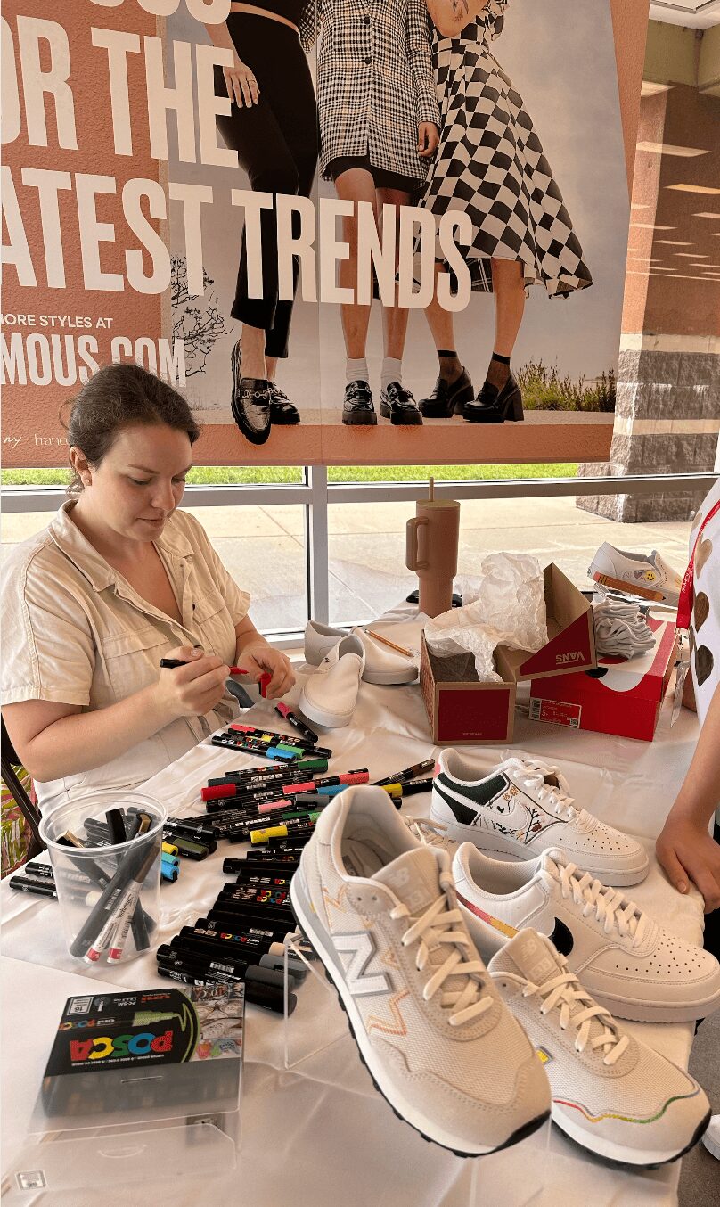 Woman sitting at a table with sneakers, makeup, and a red box, in front of a window with a poster of children in fashionable clothing.