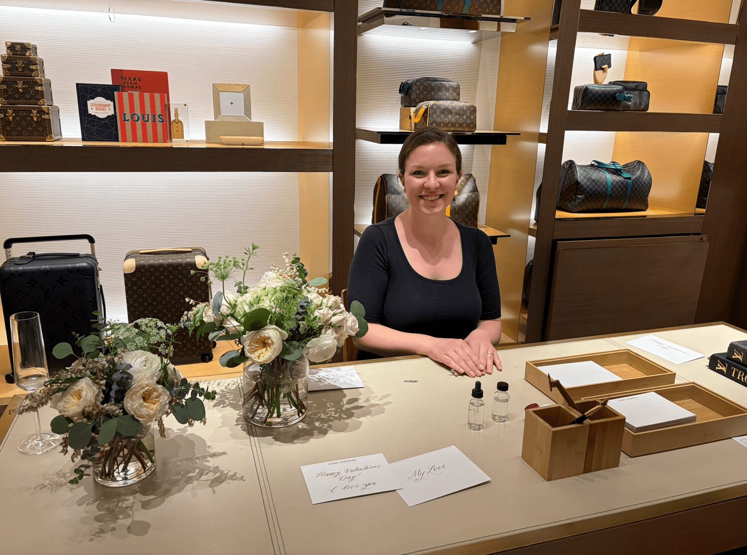 Smiling woman behind a counter with flowers, papers, and small bottles, in a store with shelves of bags in the background.