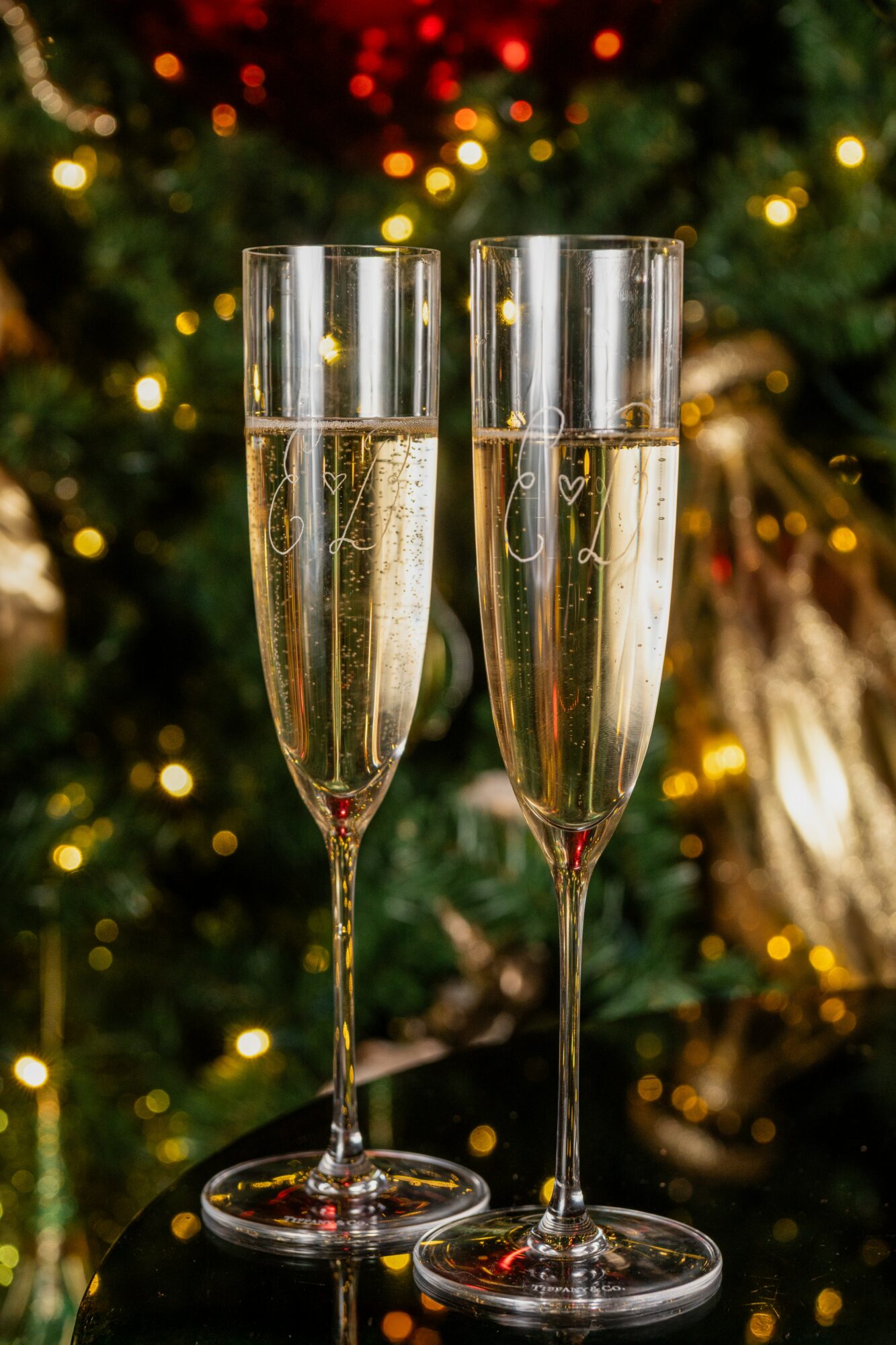 Two champagne flutes filled with sparkling wine, set against a decorated Christmas tree with lights and ornaments.