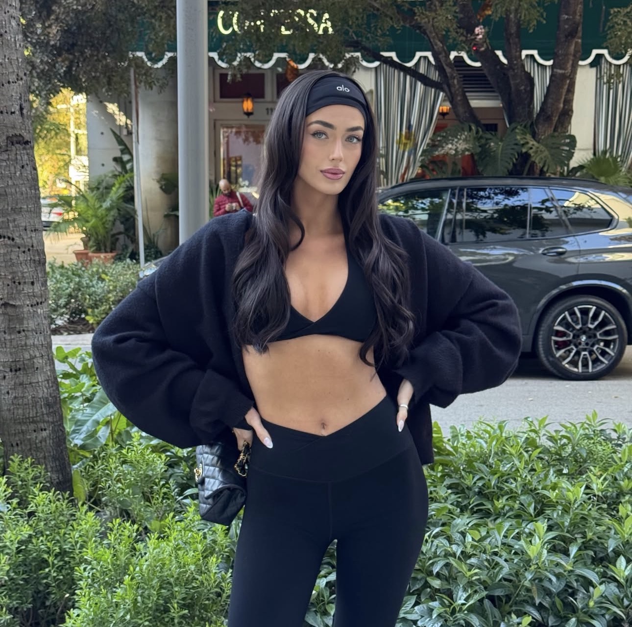 Woman with long dark hair in black sports bra and leggings, standing outdoors with greenery and a parked car behind her.