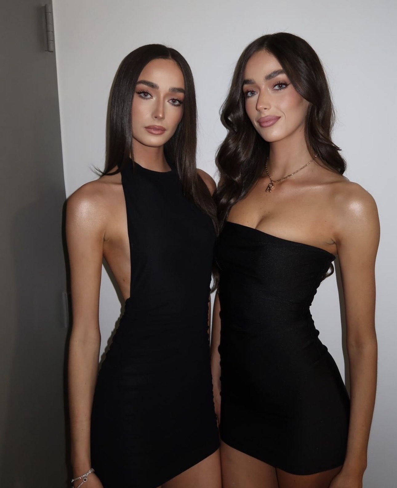 Two women with long dark hair and similar black dresses stand side by side against a plain background.