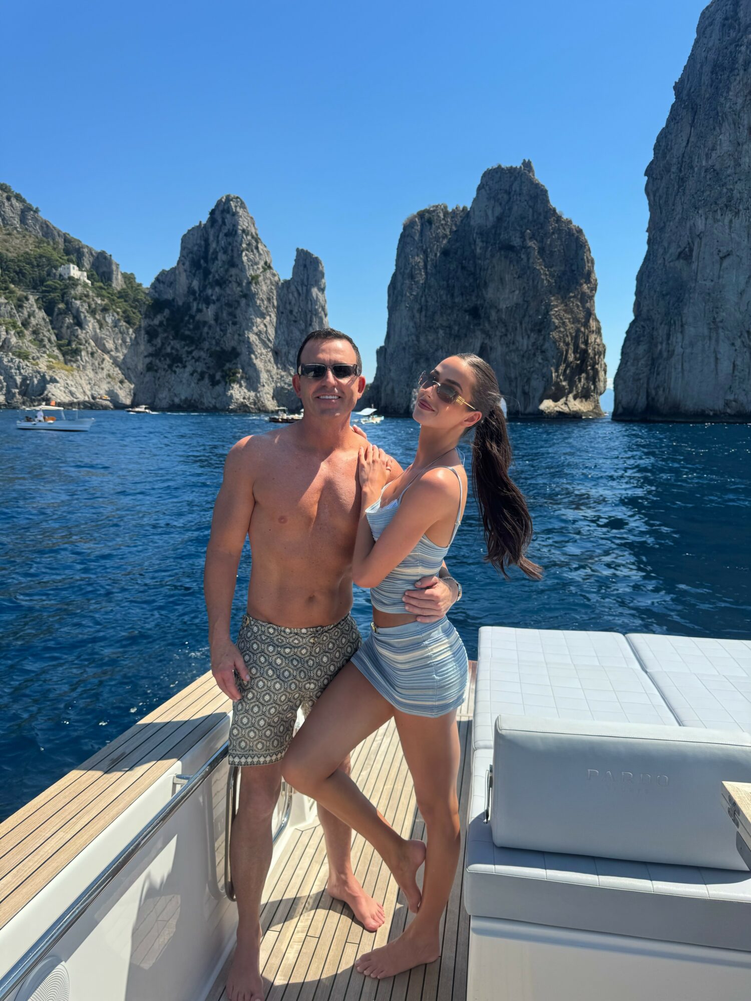 A man and woman stand on a boat with rocky cliffs and blue sky in the background.