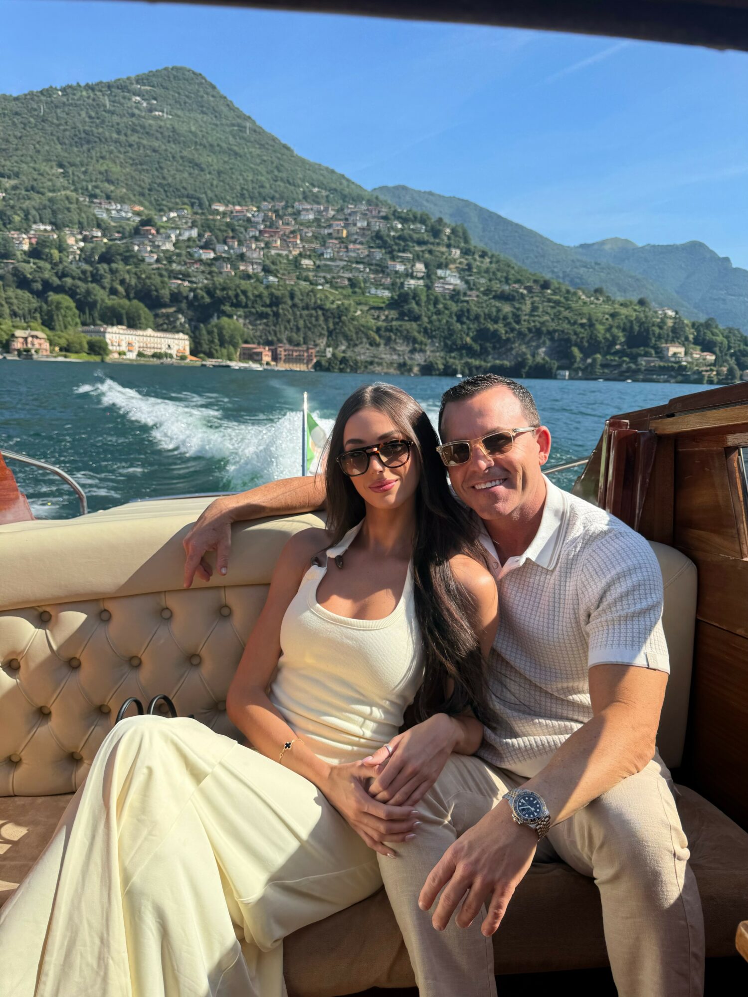 A man and woman sit together on a boat with a lake and green mountains in the background, smiling at the camera.