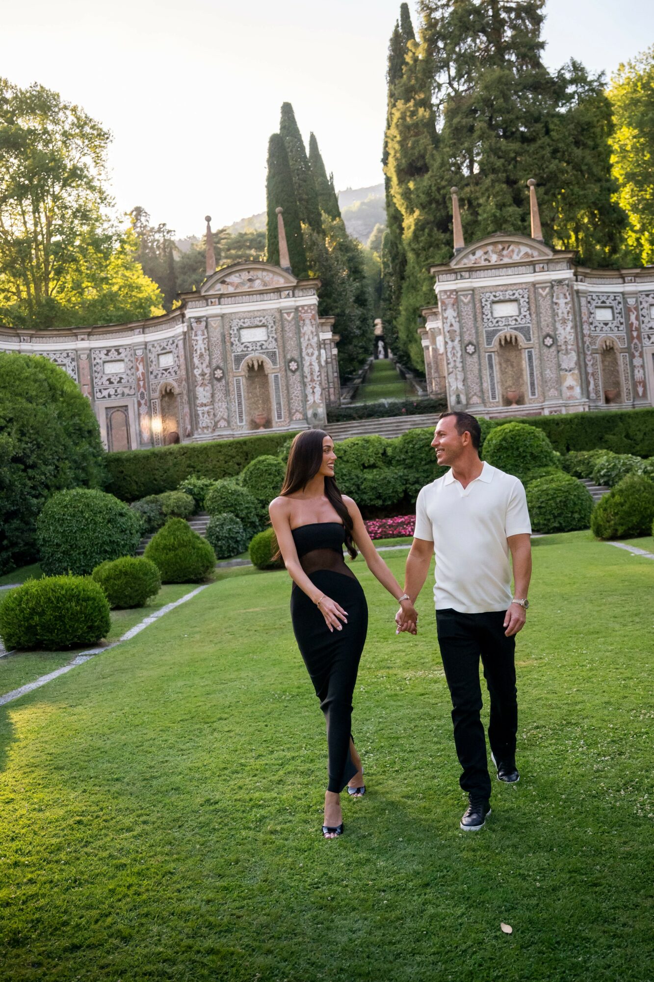 A couple holding hands walking on a green lawn with ornate stone structures and tall trees in the background.