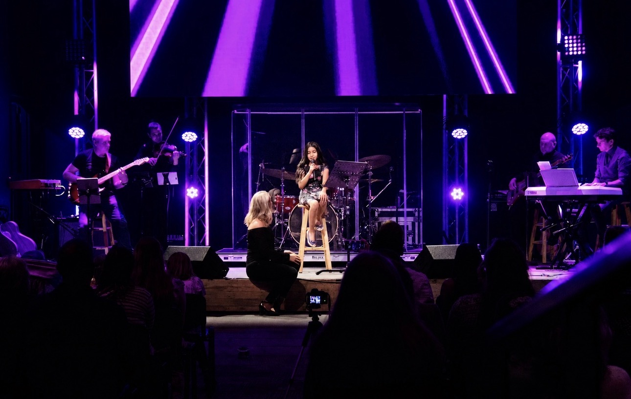 Musicians perform on stage with purple lighting, a singer in the center, and an audience watching.