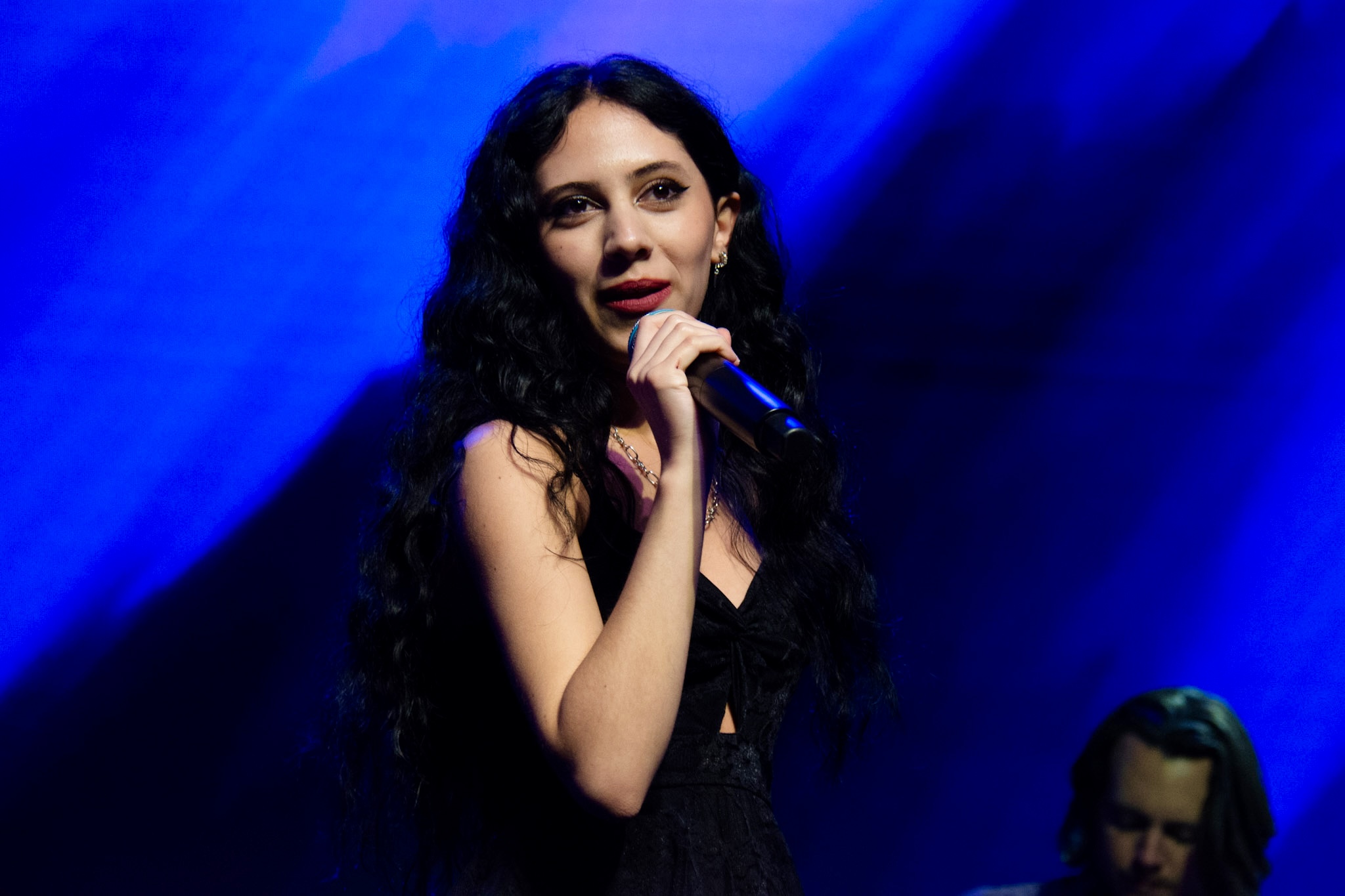 A woman with long dark curly hair holds a microphone on stage with blue lighting.