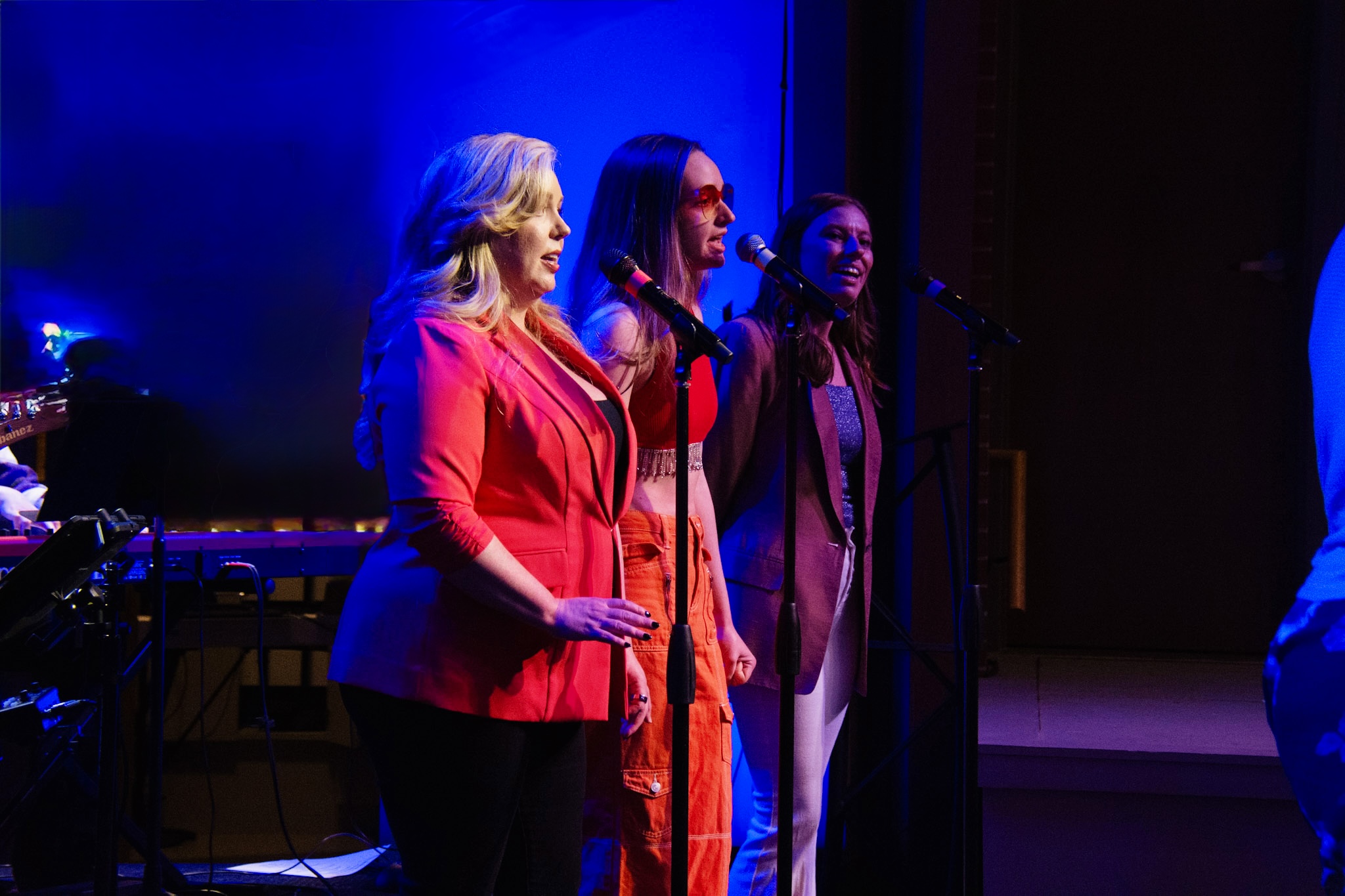 Three women sing into microphones on stage with blue and purple lighting, one wearing a red blazer, others in dresses.