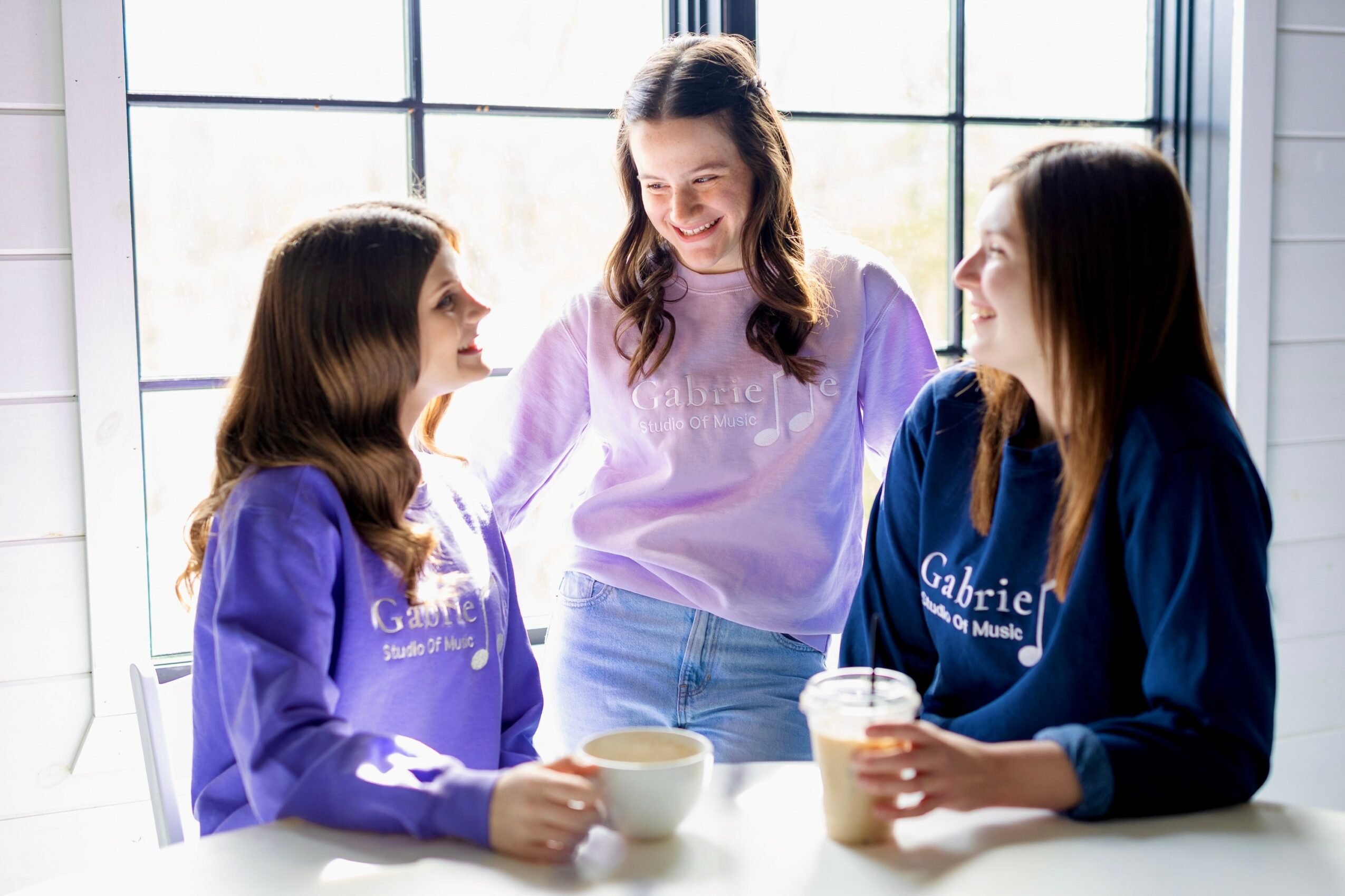 Three young women sit at a table near a large window, smiling and talking, with two holding cups and one with a drink.