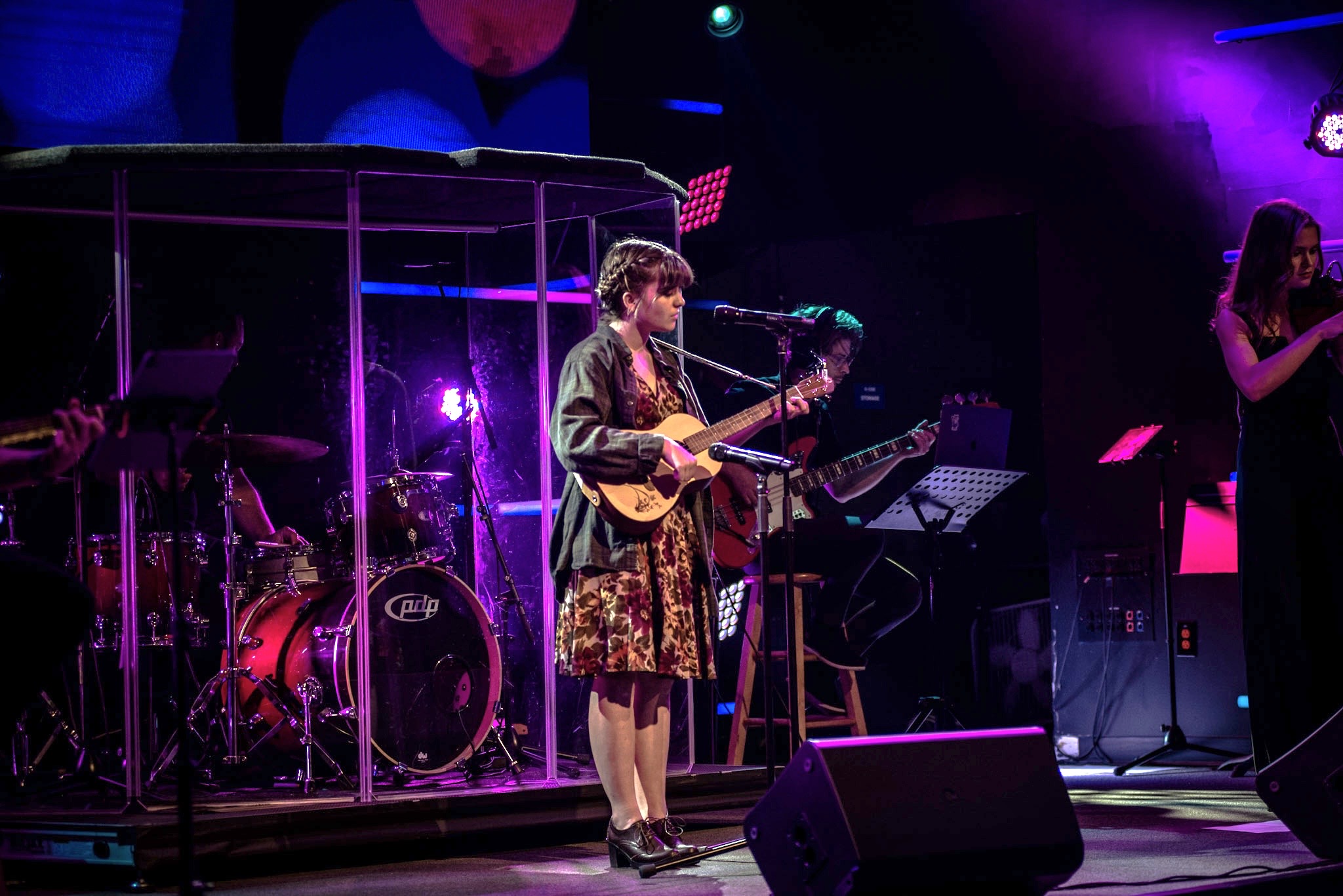 Child playing guitar on stage with band, purple lighting, and a woman singing, in a concert setting.