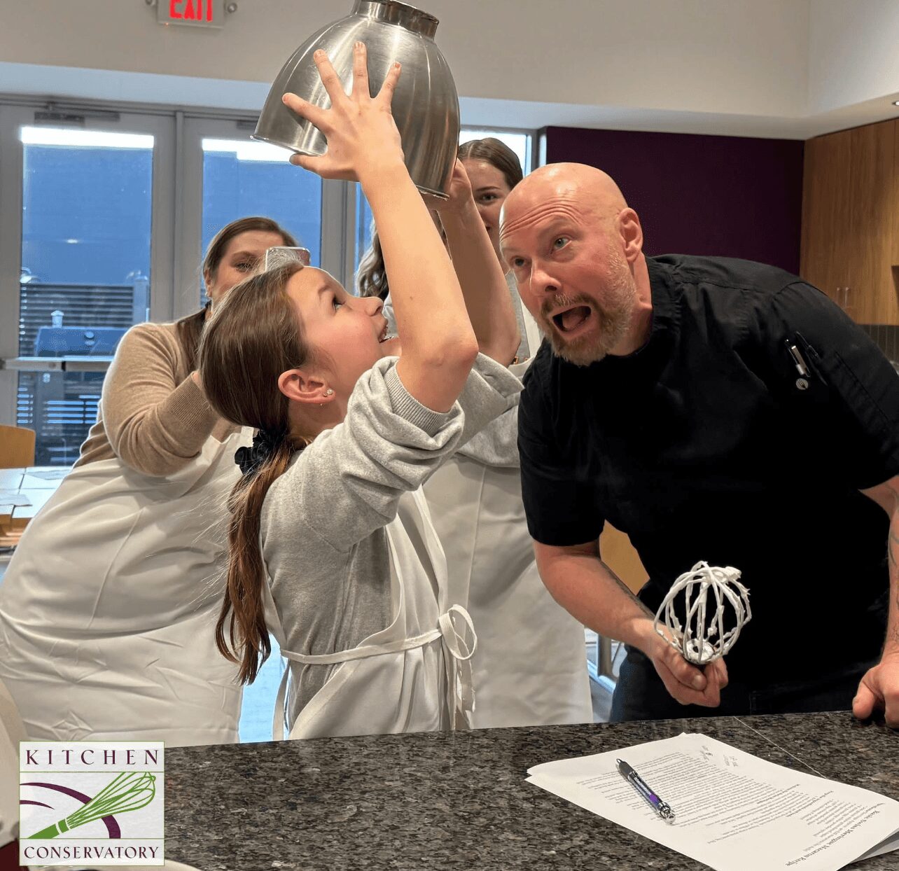 Children and adult in a kitchen, with children holding a metal mixing bowl, adult holding a whisk, all smiling.