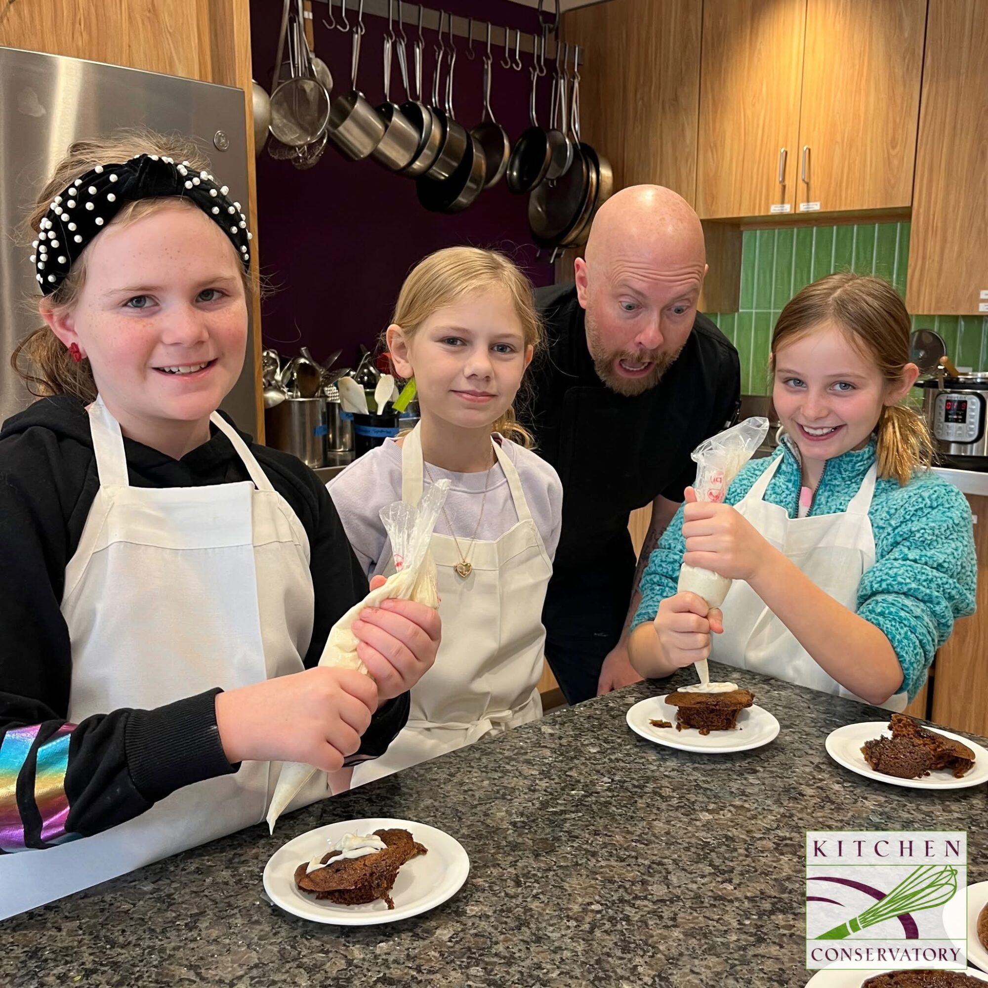 Three children and an adult in a kitchen, holding utensils and smiling, with plates of cake on the counter.