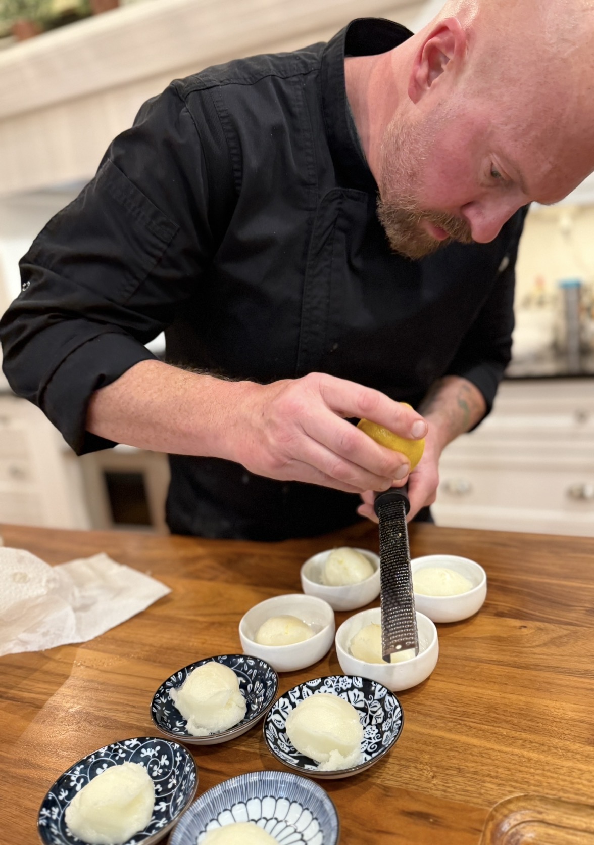 Bald man in black shirt preparing food with bowls and a knife on a wooden table.