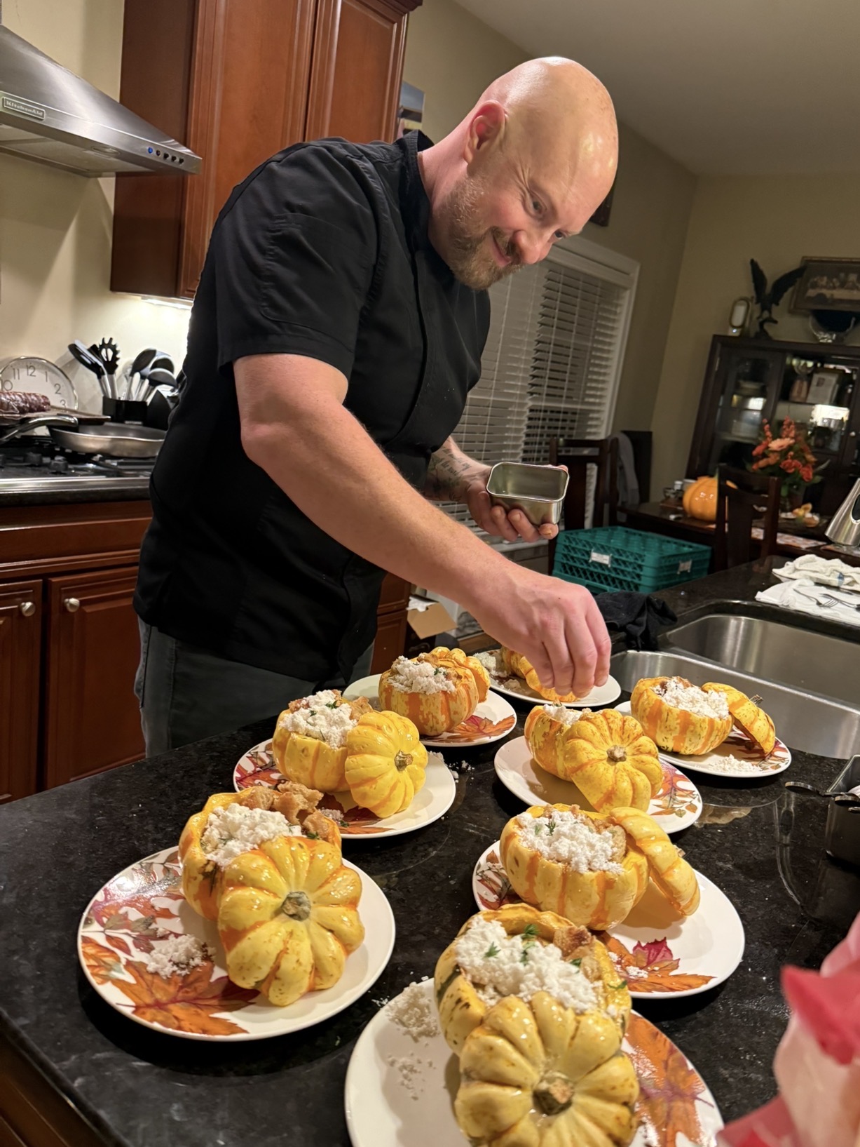 Bald man in black shirt decorating cupcakes on a kitchen counter with plates of cupcakes in front.