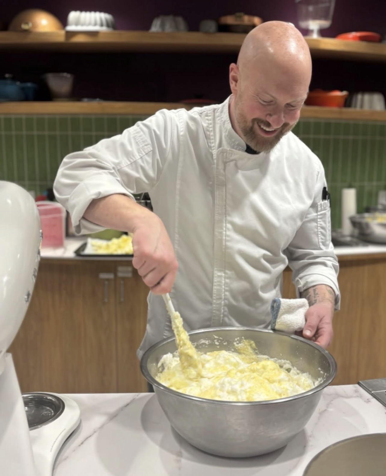 Chef stirring a large bowl of yellow mixture in a kitchen, smiling, wearing a white chef's coat.