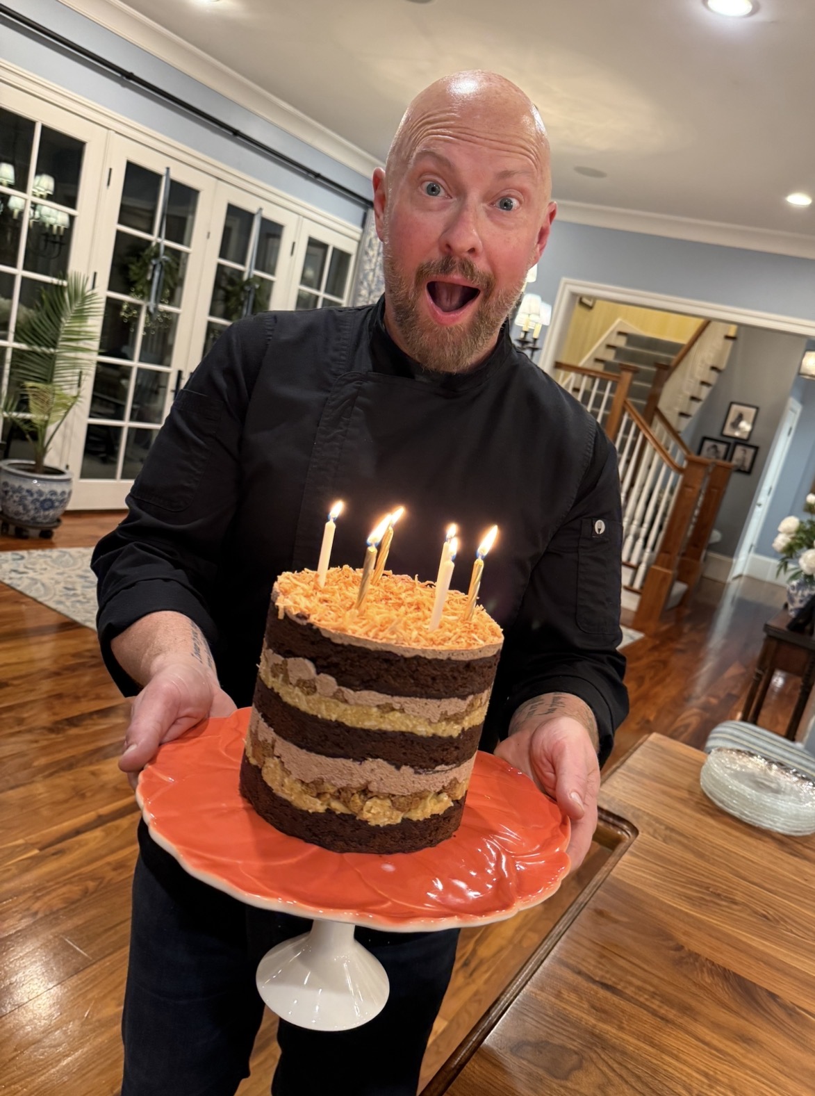 Man with a surprised expression holding a layered birthday cake with lit candles on a red cake stand in a dining room.