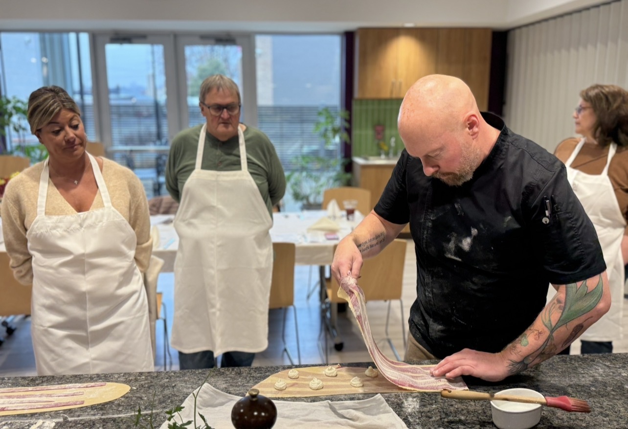 Chef demonstrating bacon wrapping to group of people in a kitchen, with large windows in the background.