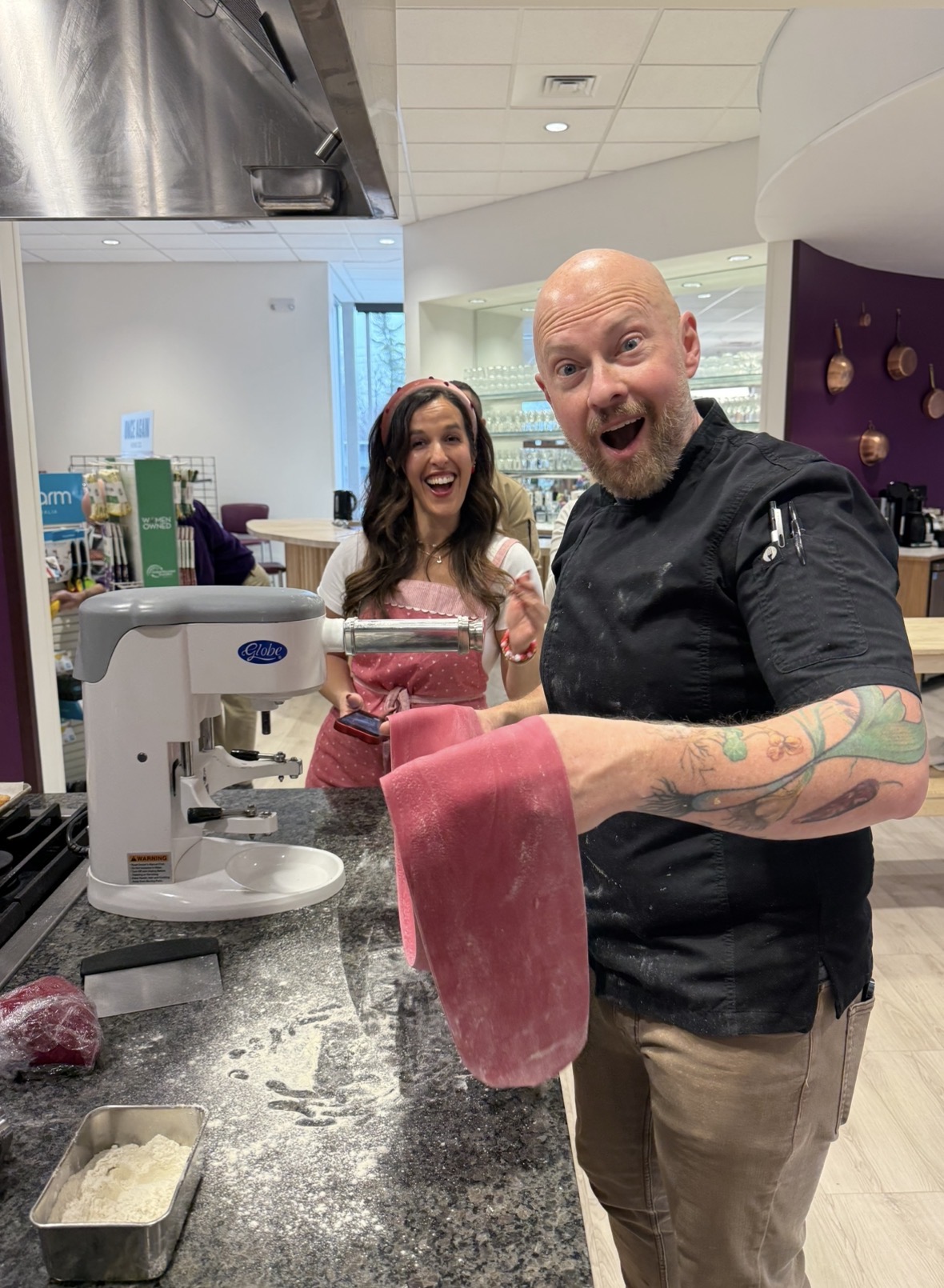Two people smiling in a modern kitchen, one holding a pink towel, with a scale and kitchen items nearby.