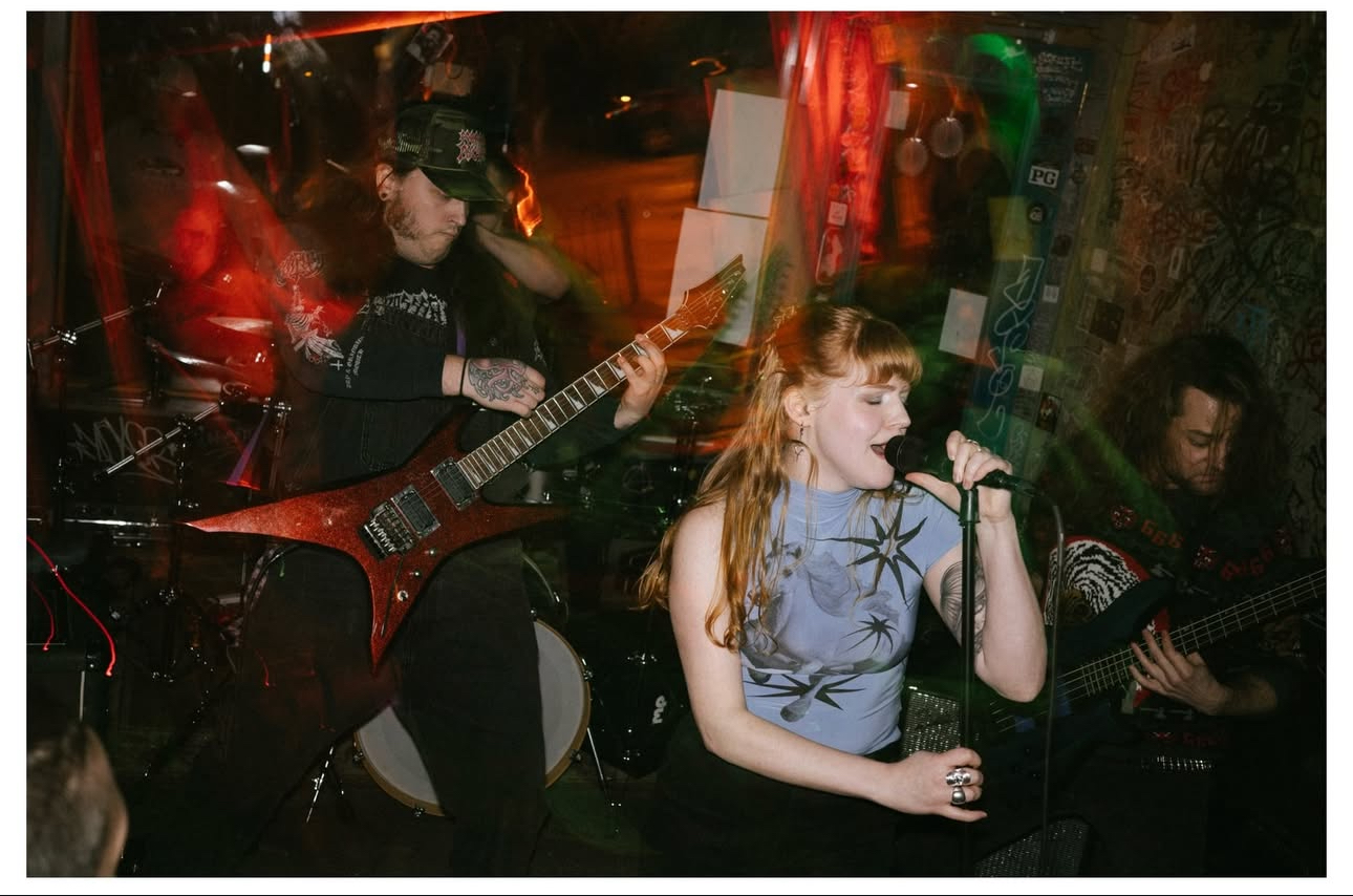 Young woman singing into a microphone with two guitarists playing beside her in a dimly lit setting.