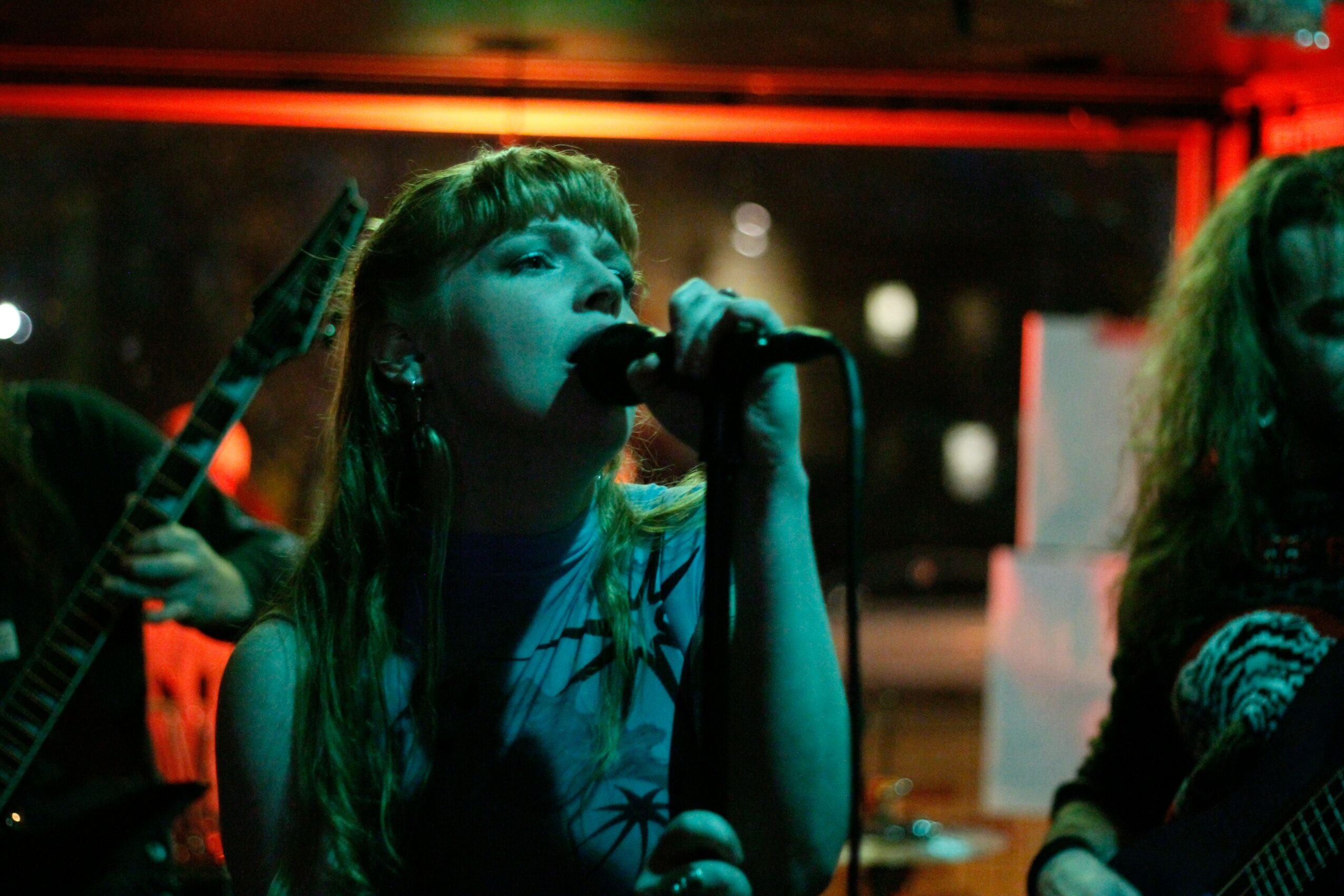 Young woman singing into a microphone on stage with red and green lighting, holding a guitar, with another person partially visible.