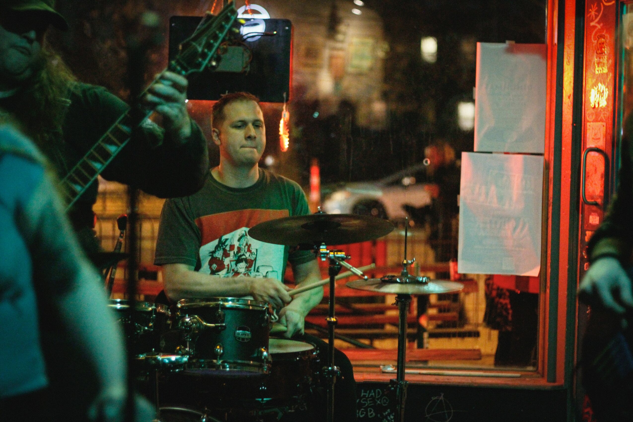 Musician playing drums and another playing guitar in a dimly lit indoor setting, with a window showing streetlights outside.