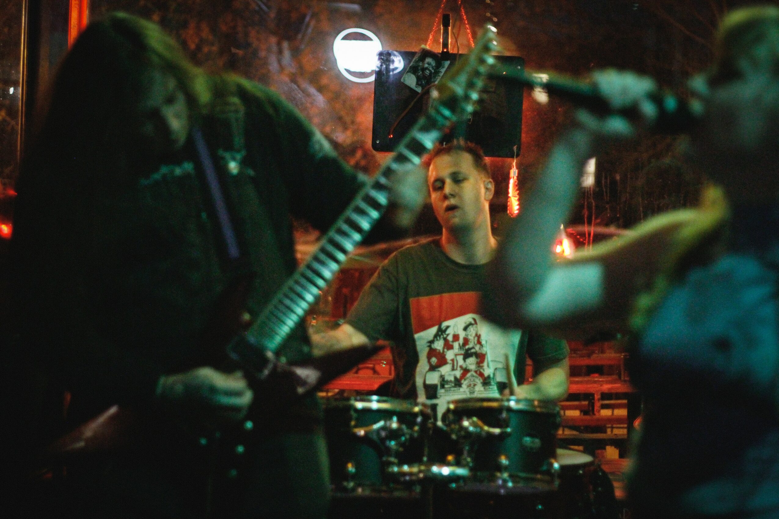 Three people playing musical instruments in a dimly lit room, with a person in the center looking at the guitarist.