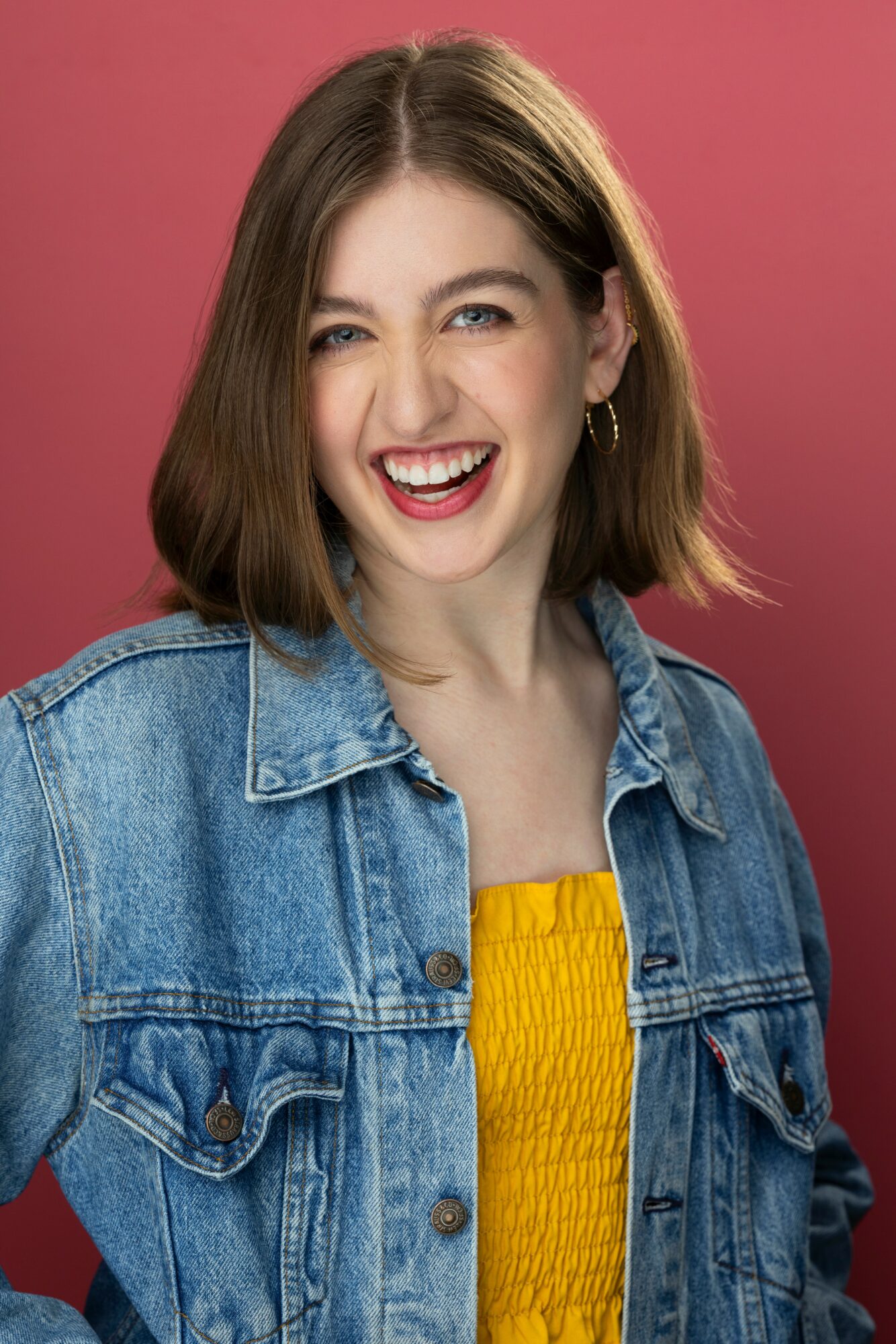 Young woman with shoulder-length brown hair, smiling and winking, wearing a denim jacket and yellow top, pink background.