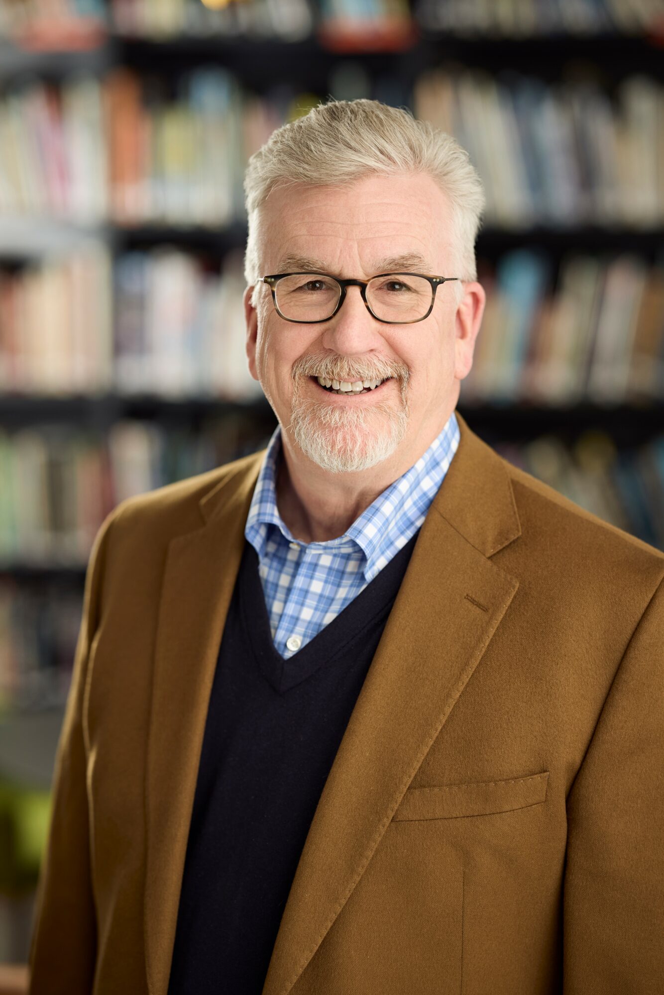 Smiling older man with glasses, gray hair, and beard, wearing a brown blazer and blue checkered shirt, in a library.