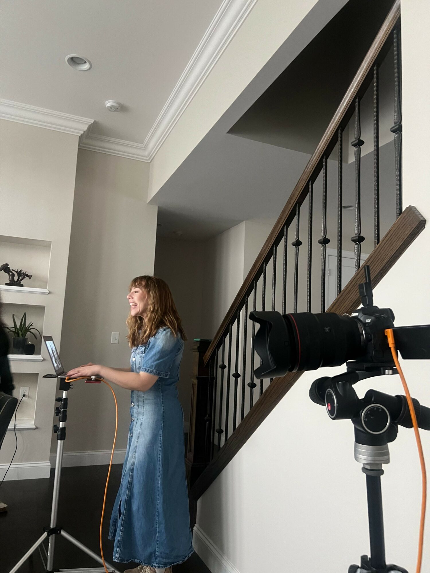 Woman in a blue dress standing in a room with a staircase and camera equipment, holding a tablet or phone.