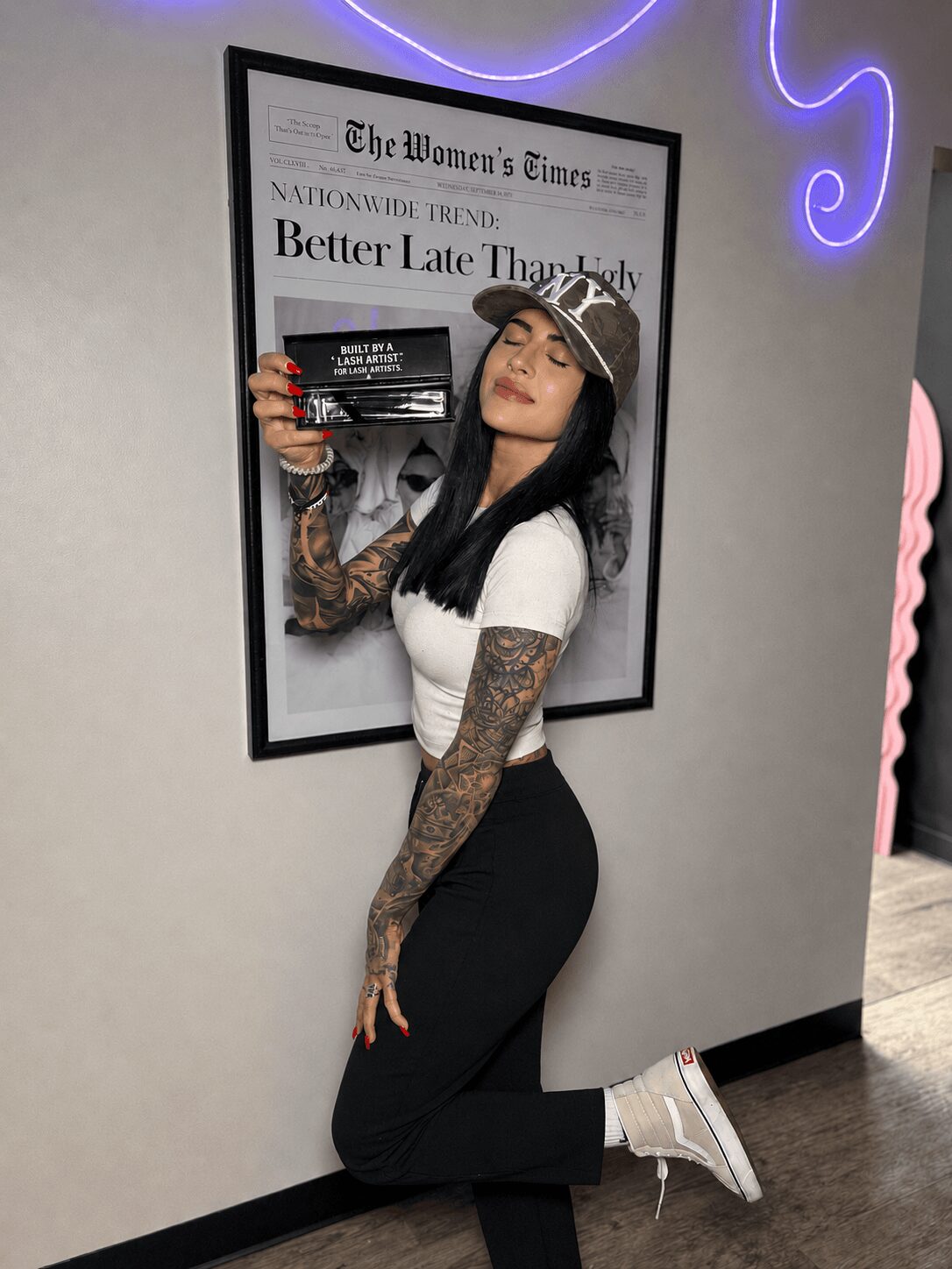 Young woman with tattoos, wearing a white top, black pants, and sneakers, taking a mirror selfie in front of a framed newspaper.