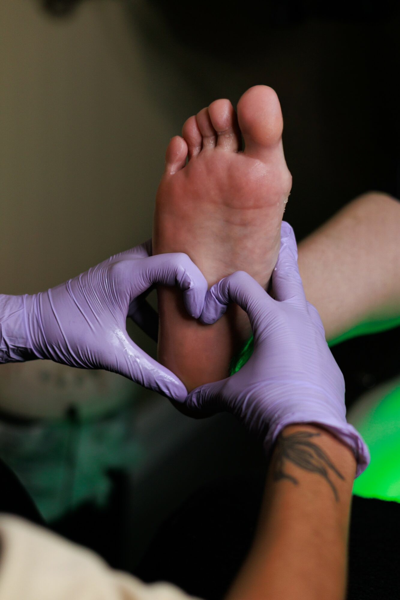 Person's foot held by gloved hands, forming a heart shape with fingers, with a tattooed arm visible.