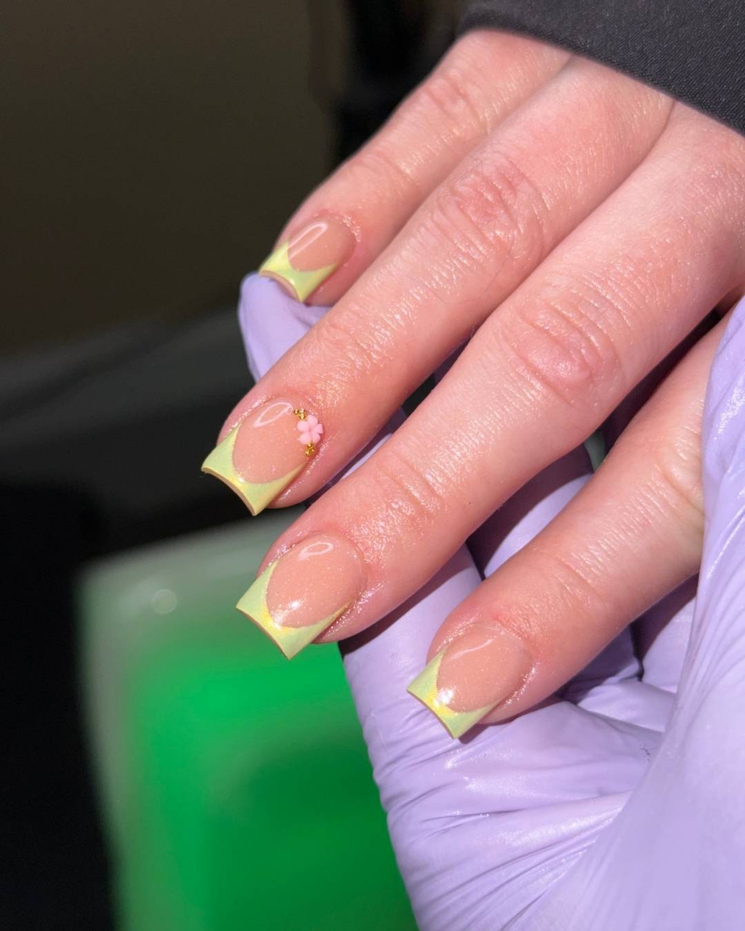 Hand with manicured nails, showing yellow tips and pink accents, held by a gloved hand.