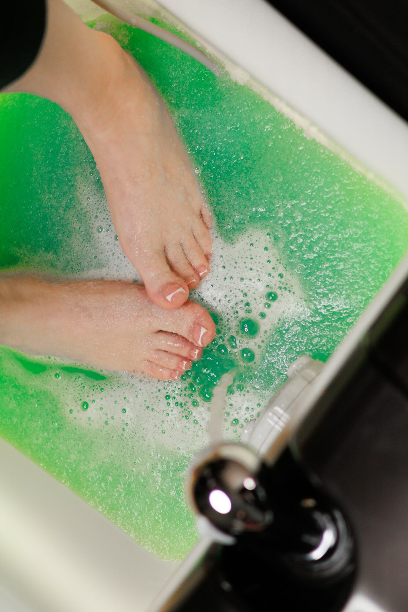 Person's feet in a green bathtub filled with soapy water, with bubbles and foam visible.