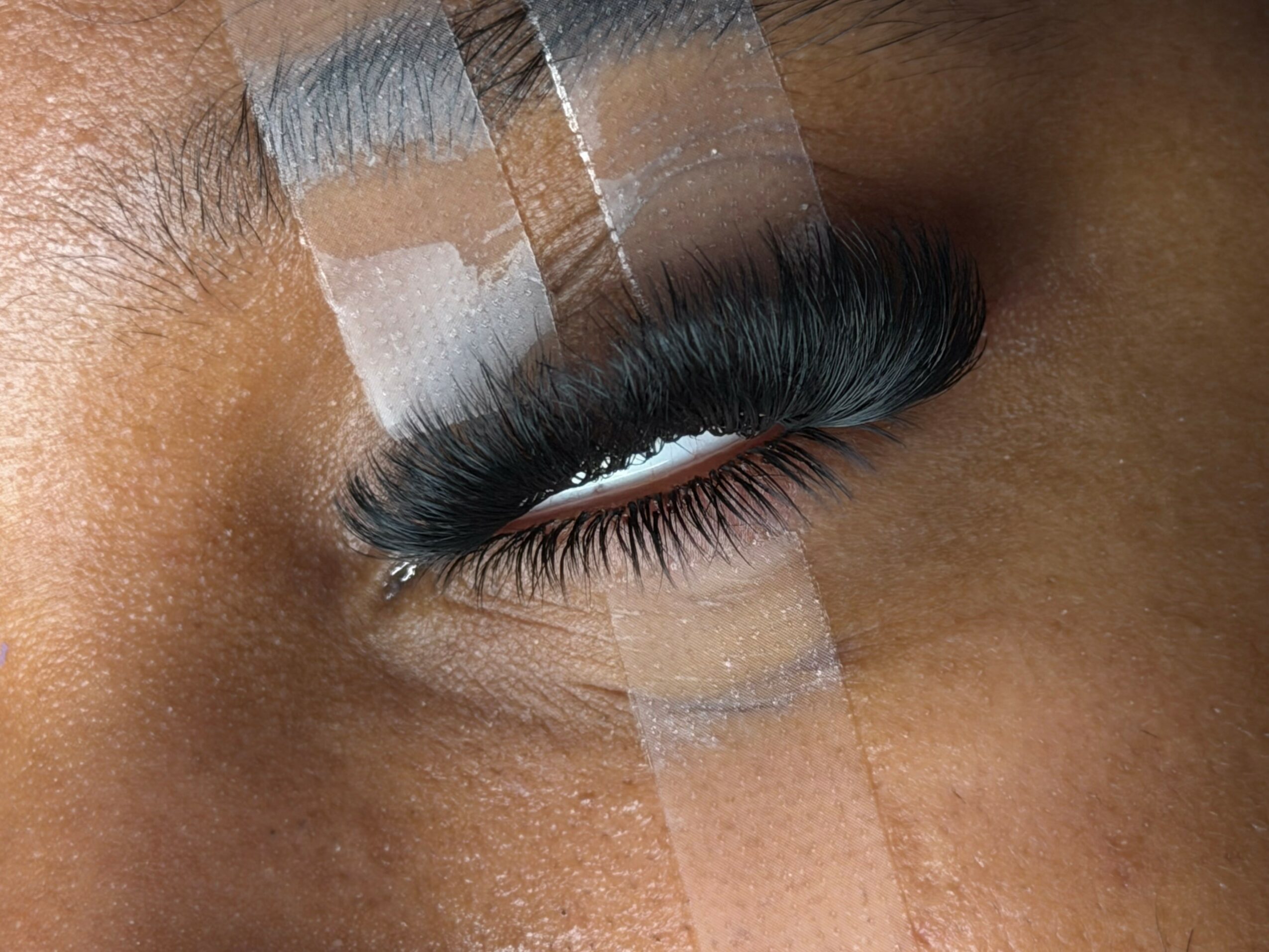Close-up of a person's eye with long, dark eyelashes and clear tape on eyelid, skin visible around eye.