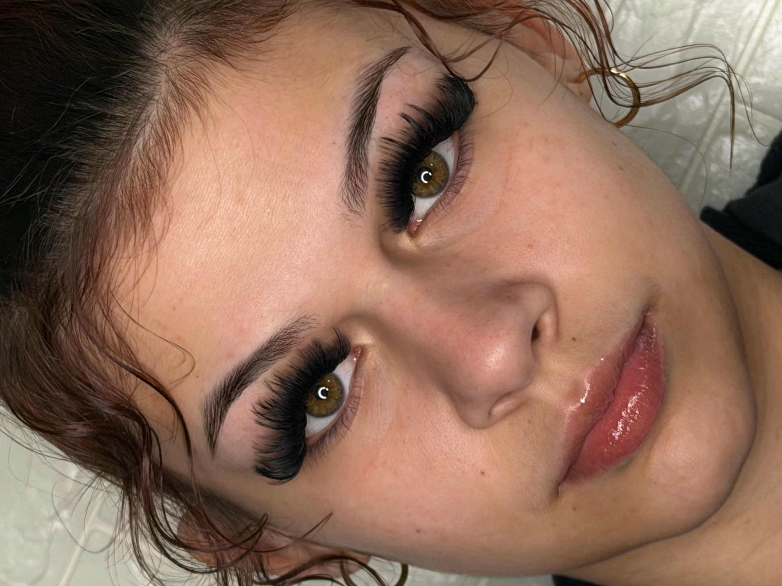 Close-up of a woman's face with makeup, lying down, looking at the camera, with curly hair and glossy lips.