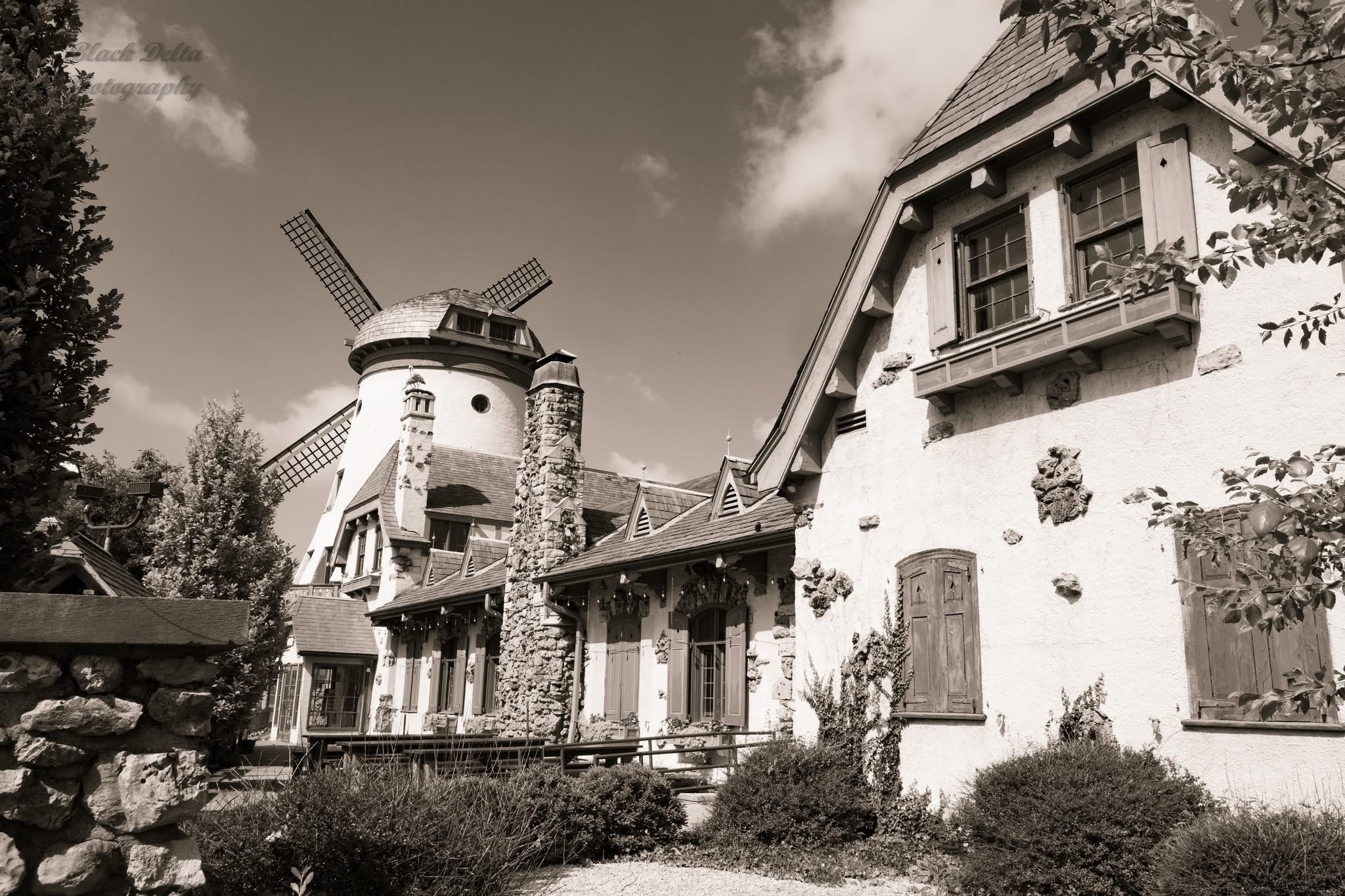 A windmill with four blades next to a house with multiple windows and a sloped roof, surrounded by trees and bushes.