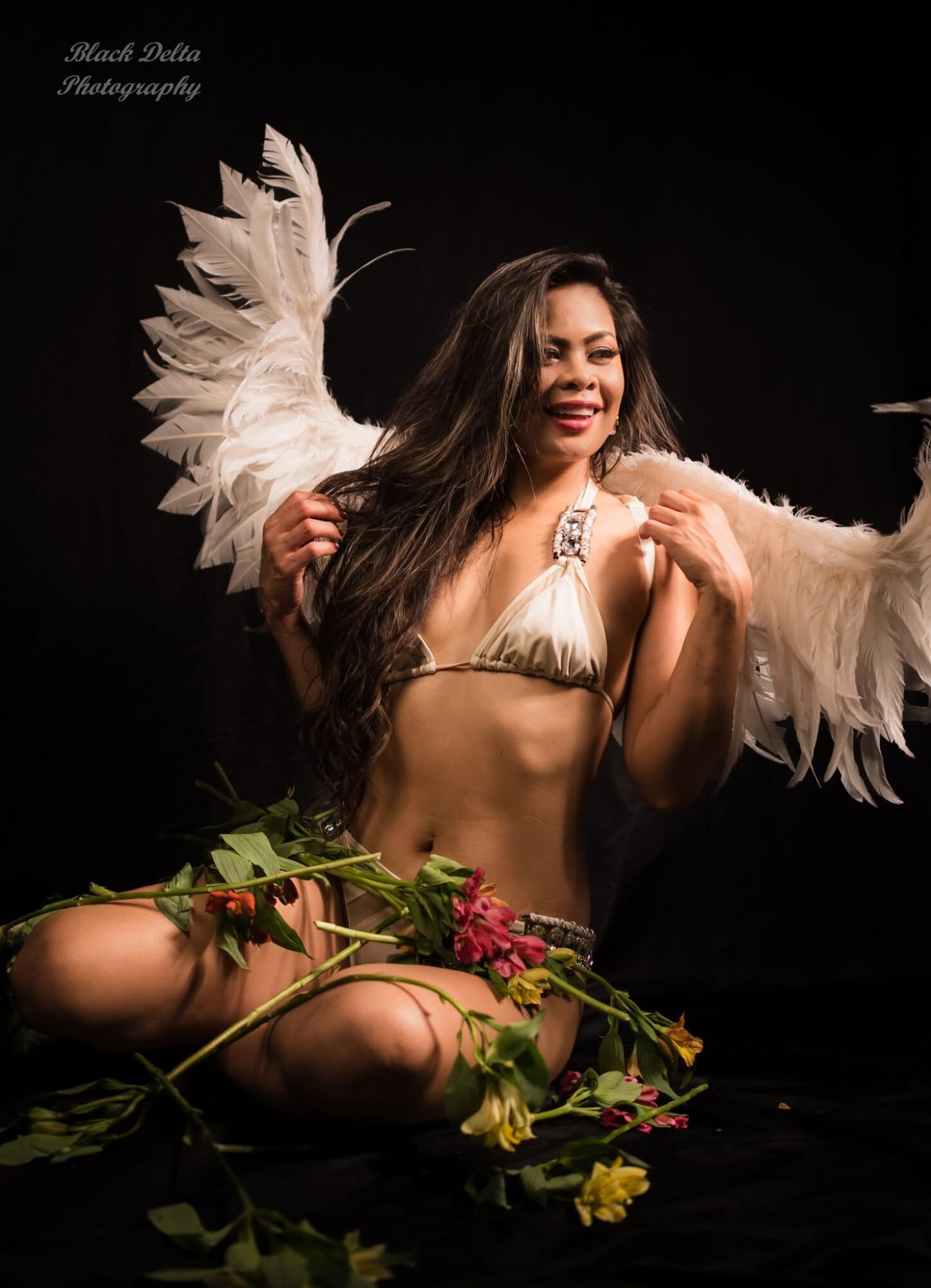Woman with long hair smiling, wearing angel wings, sitting with flowers and greenery, black background.