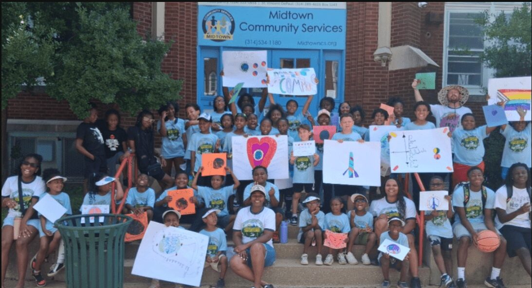 Group of children and adults holding colorful drawings outside Midtown Community Services building.