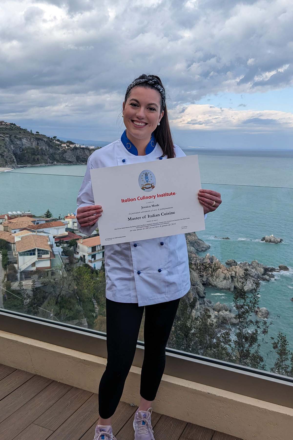 Woman in chef uniform holding certificate with coastal landscape and buildings behind her.