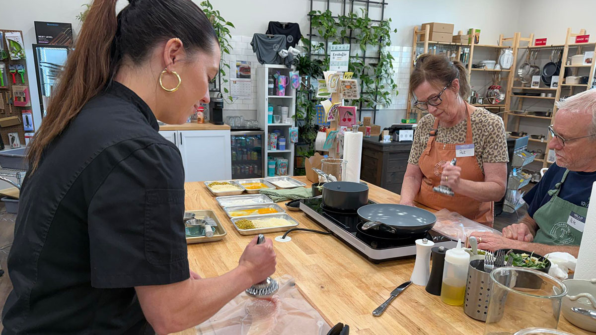 Three people in a store or workshop, one woman preparing something at a wooden table, two others observing, shelves in background.