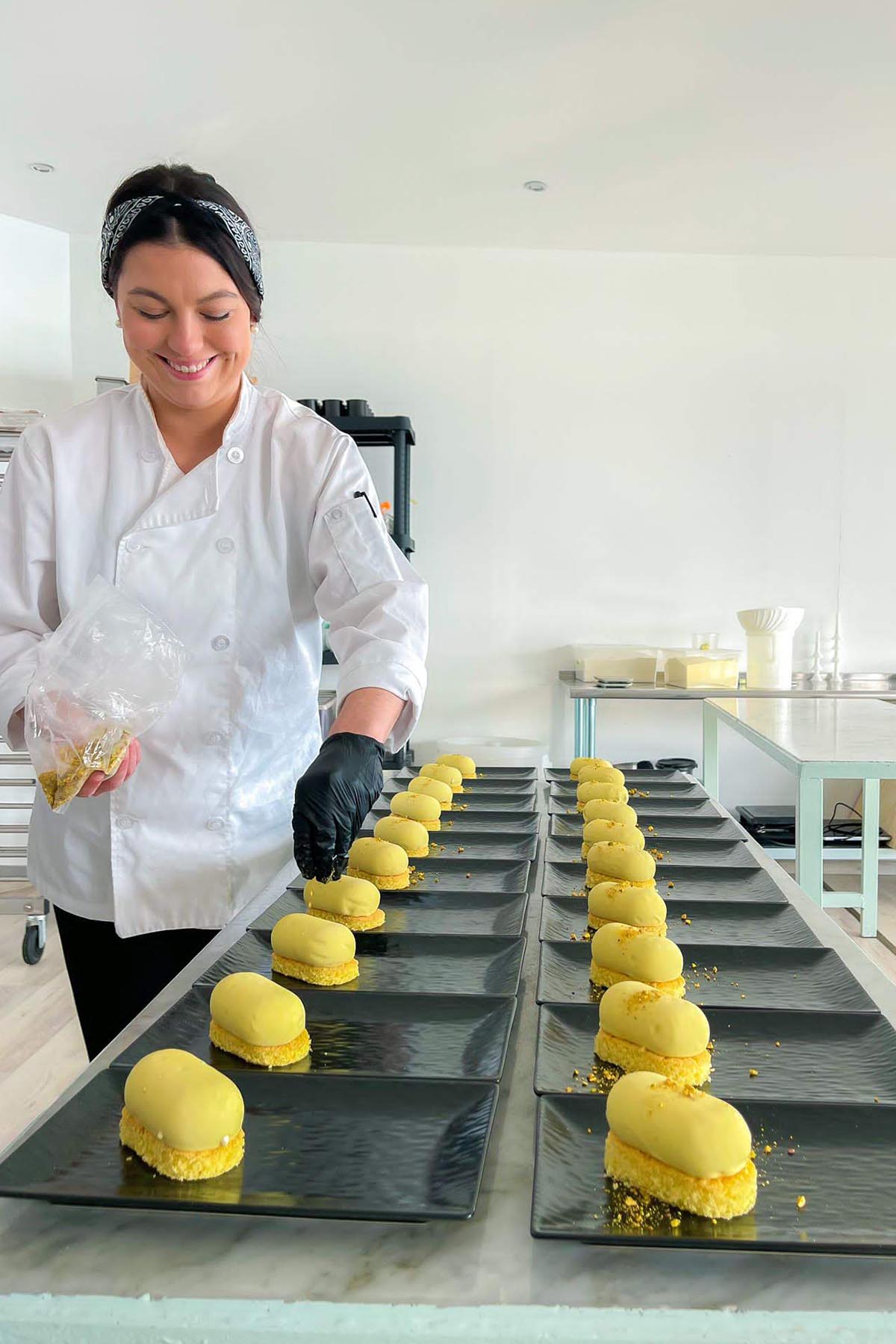 Woman in chef's coat and gloves preparing yellow desserts on black trays in a kitchen, smiling.