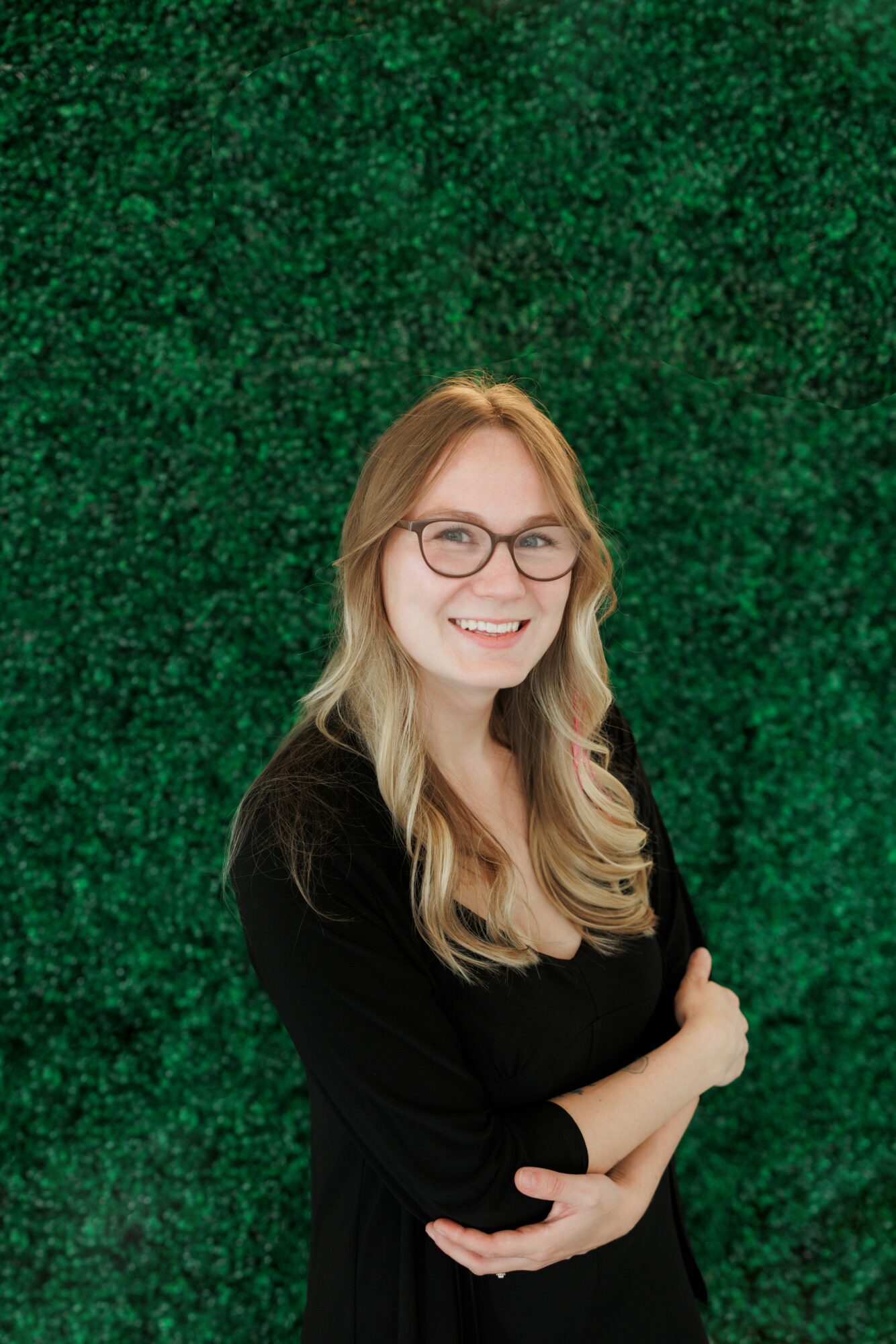 Smiling woman with glasses and long hair, wearing a black top, standing in front of a green textured background.