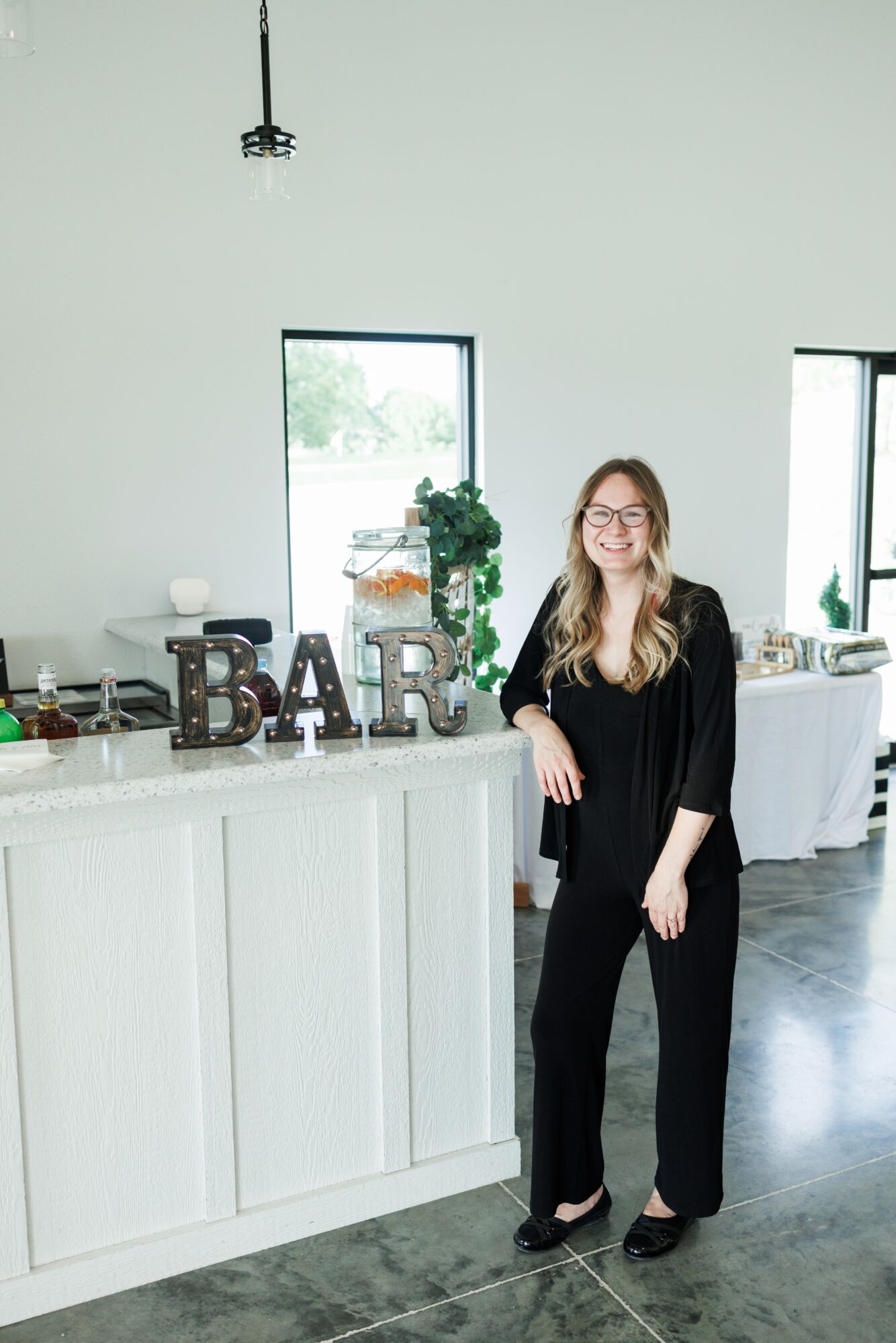 Woman with glasses and long hair standing at a bar counter in a bright room, smiling, wearing a black outfit.