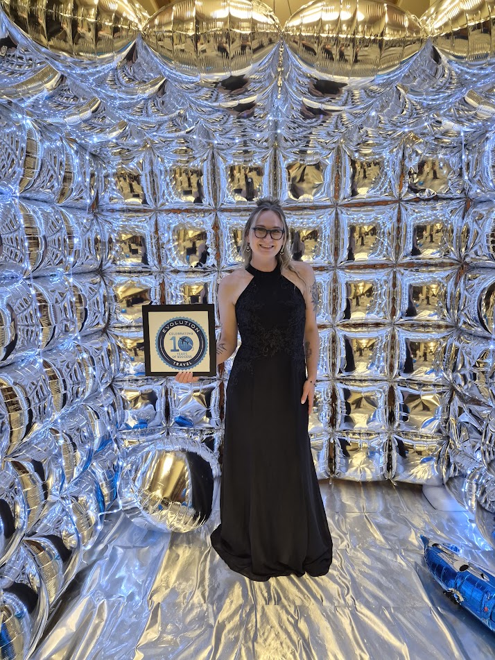 Woman in a black dress and glasses standing in front of a shiny, reflective backdrop with gold and silver balloons.